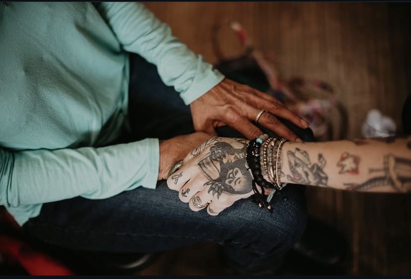 Two people hold hands, one with tattoos and bracelets, the other with a ring, in a close-up shot.