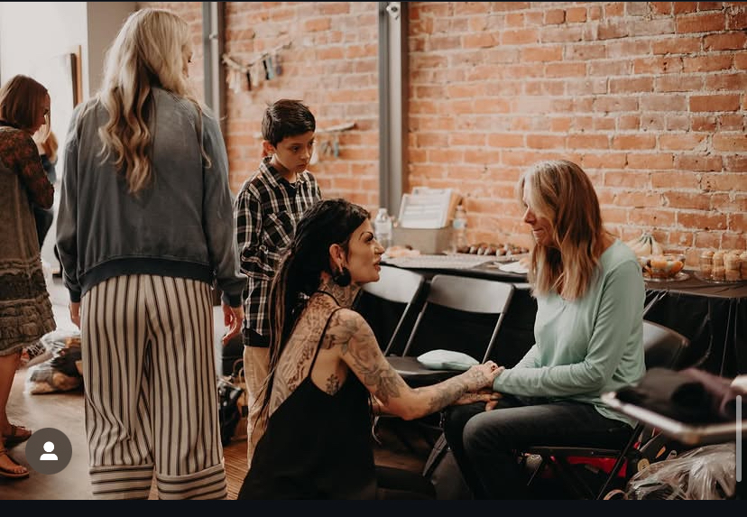 Two women sit and hold hands, engaging in conversation, with three children standing nearby in a brick-walled room.