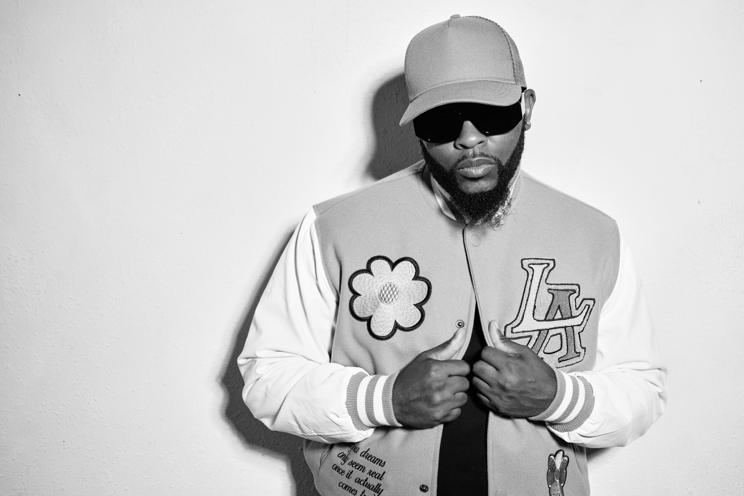 Black and white photo of a man wearing a cap, sunglasses, and a varsity jacket with a flower patch and LA logo.