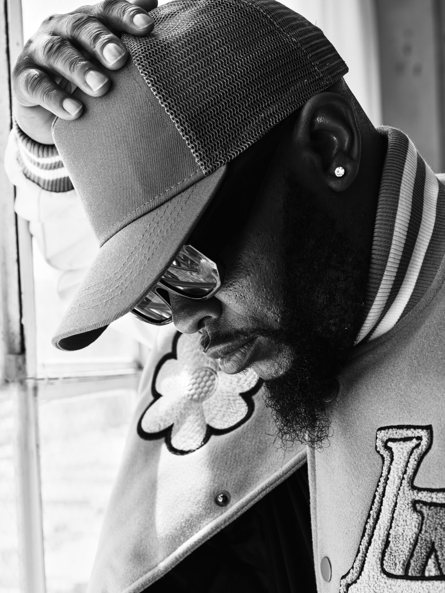 Man wearing a cap and sunglasses, touching his head, with a jacket featuring a letter patch, in black and white.