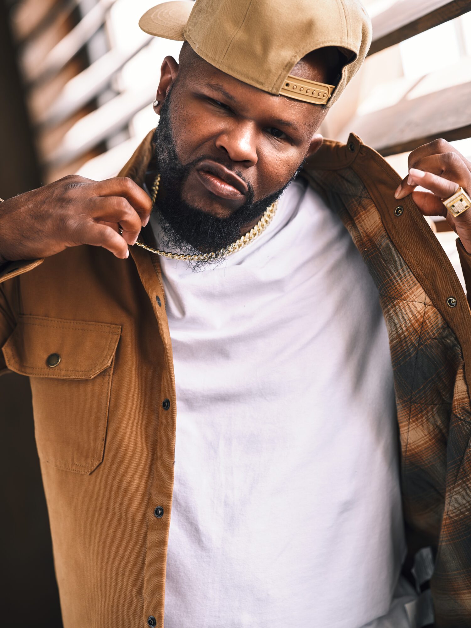Man with a beard wearing a tan cap and jacket, adjusting his jacket, outdoors.