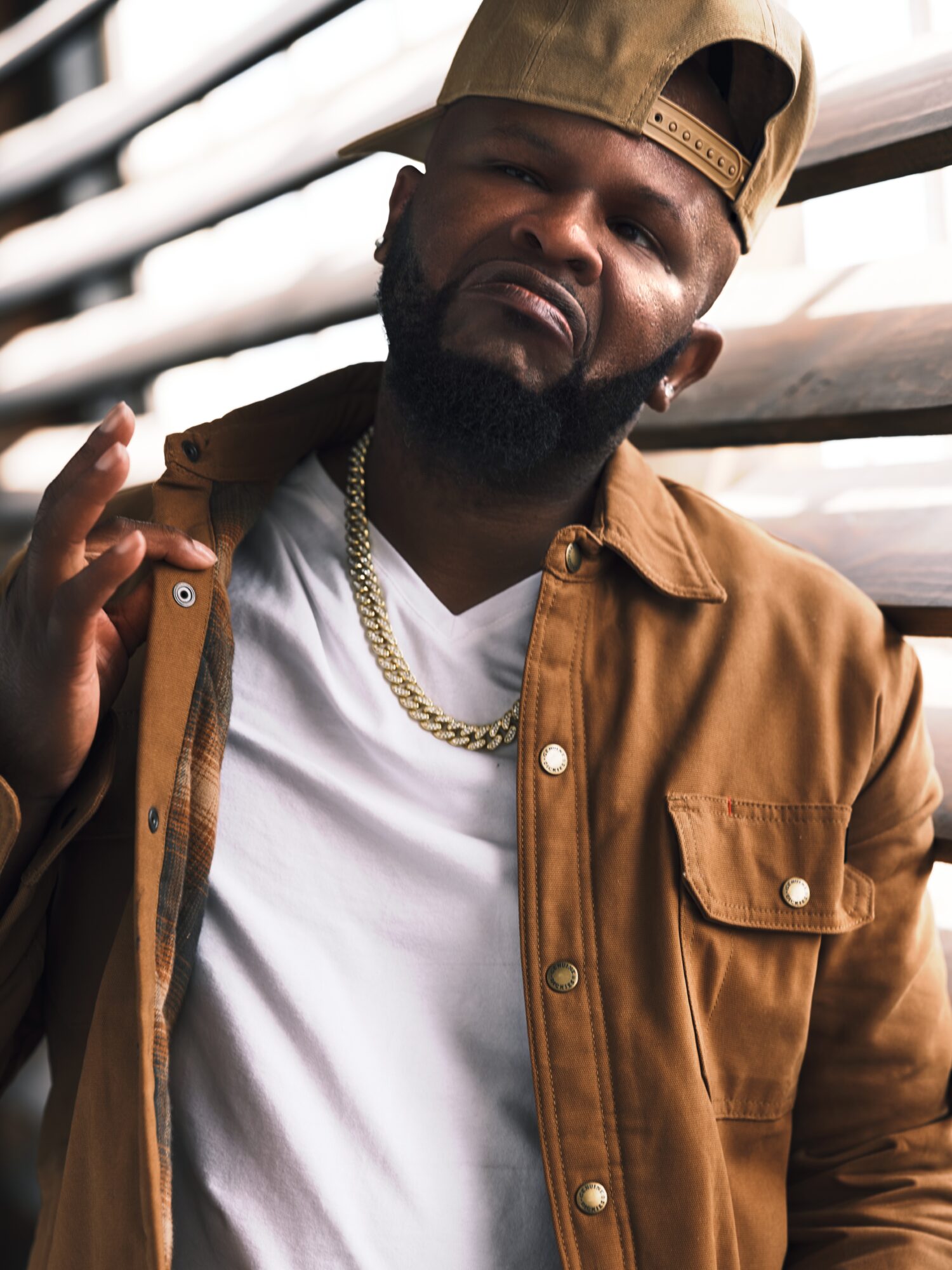 Man with a beard wearing a tan cap, brown jacket, white shirt, and gold chain, making a hand gesture.