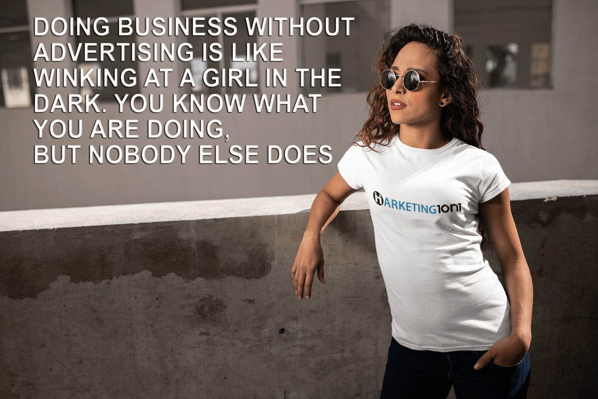 Pregnant woman wearing sunglasses and a white T-shirt with text, standing outdoors against a wall, smiling.