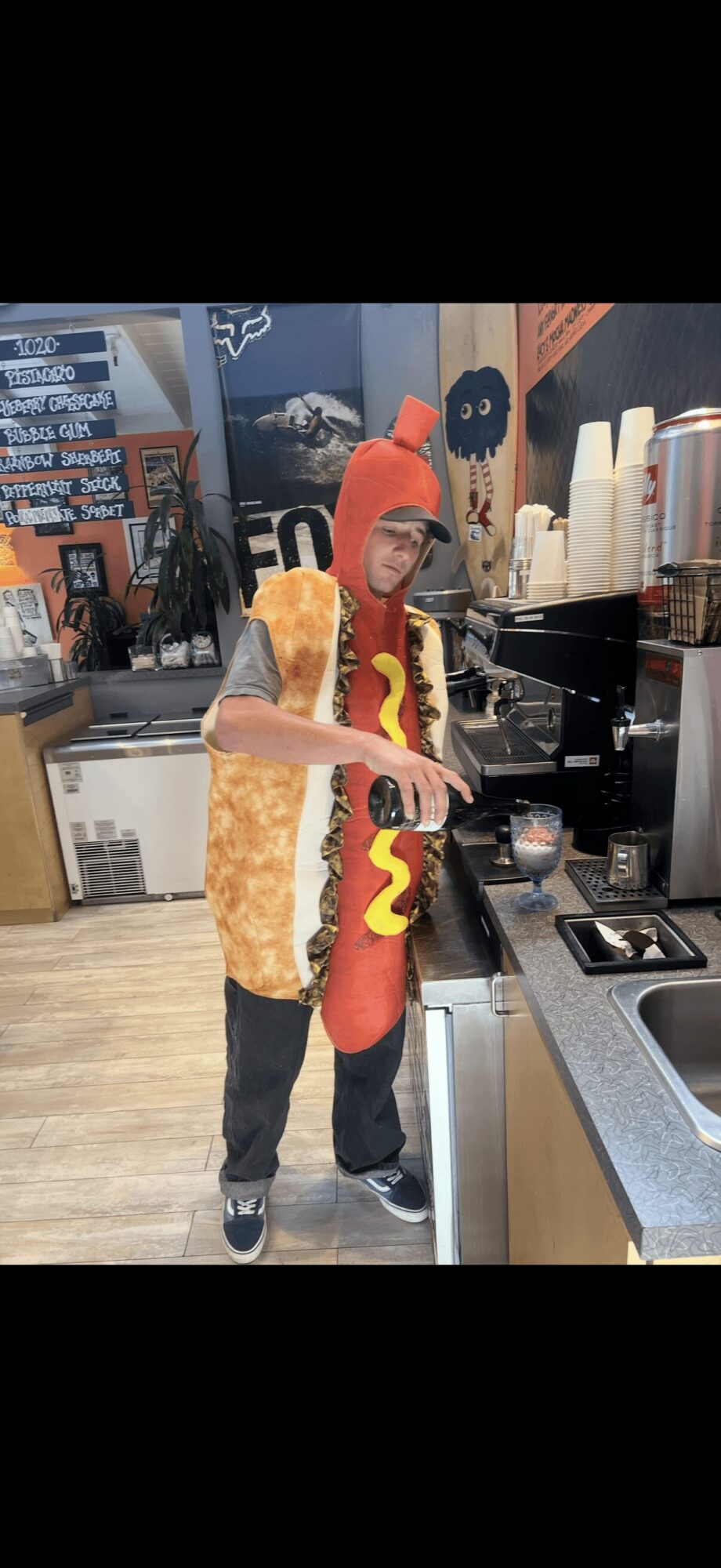 Person dressed in a hot dog costume preparing a drink at a counter in a store or cafe.