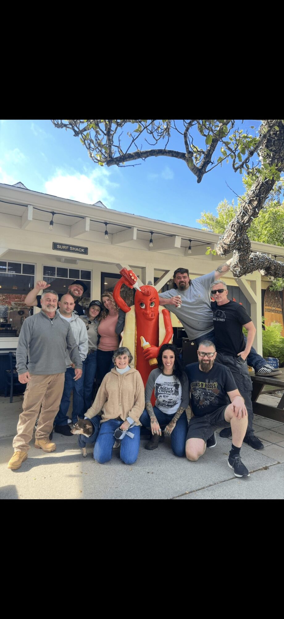 Group of people posing outdoors with a person in a red costume, in front of a building and tree, sunny day.