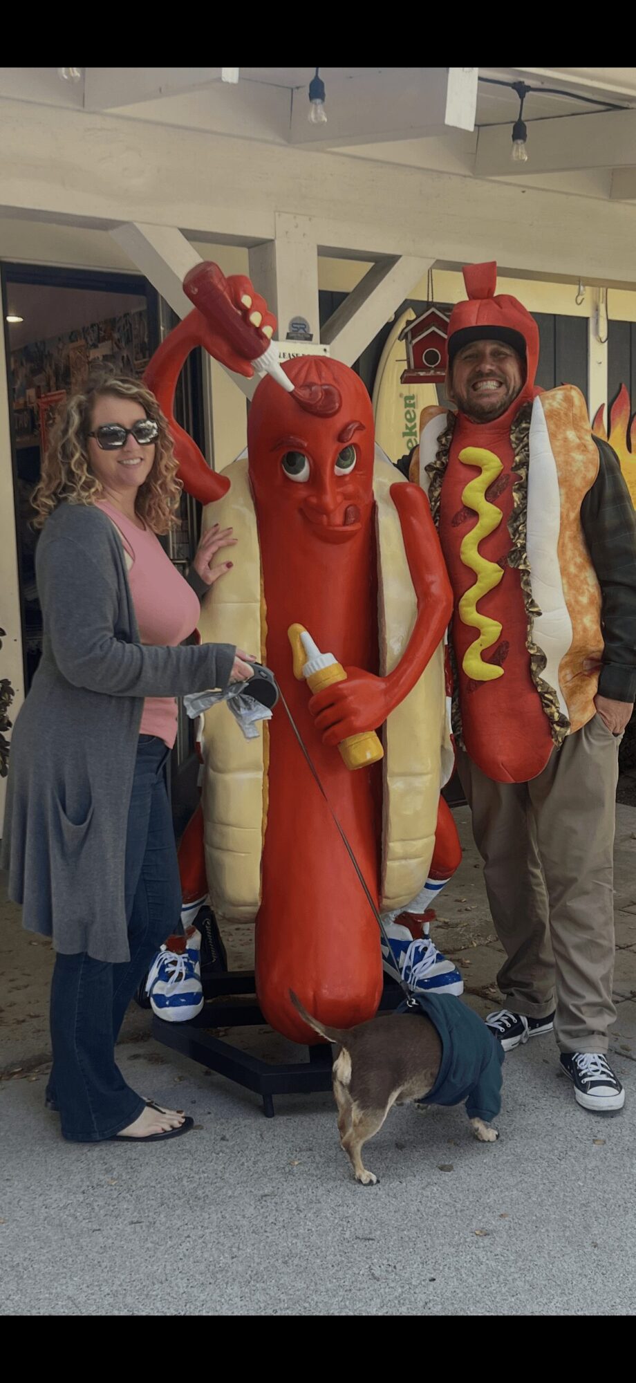 Two people stand with hot dog and ketchup bottle mascots, one dog in front, indoors, with ceiling lights and windows.