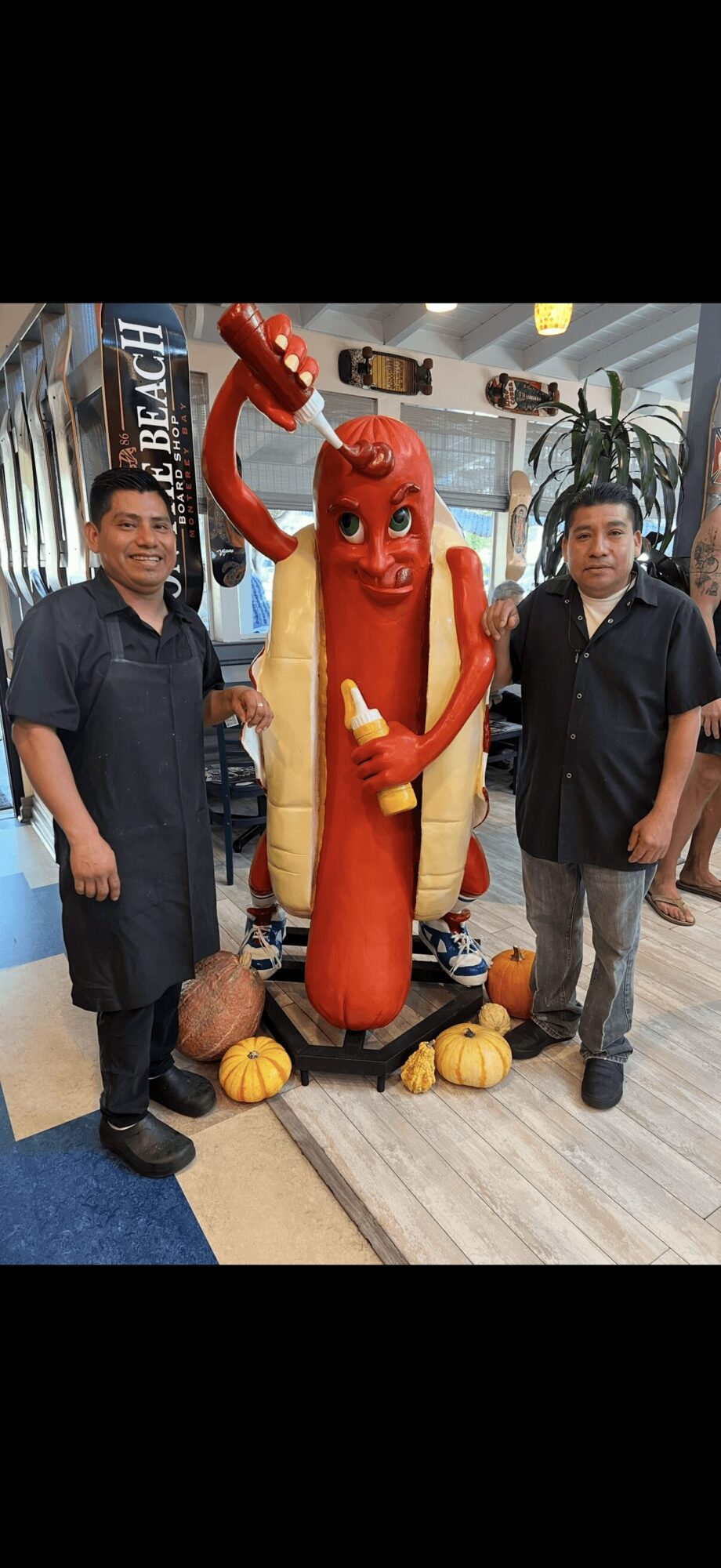 Two people stand beside a large hot dog sculpture with a red face and arms, in an indoor setting.