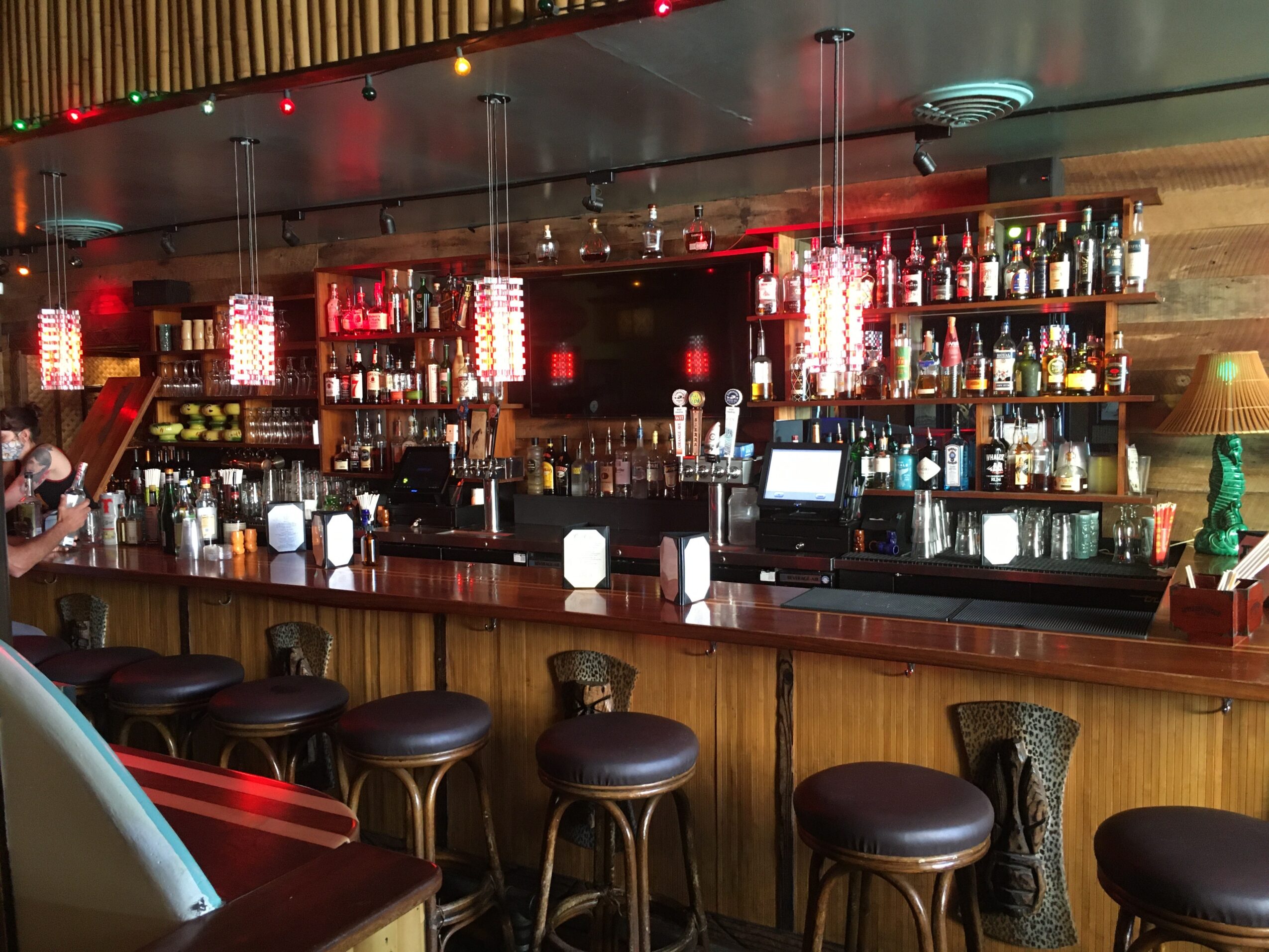 Bar with shelves of bottles, illuminated by hanging lights, and a row of stools in front.