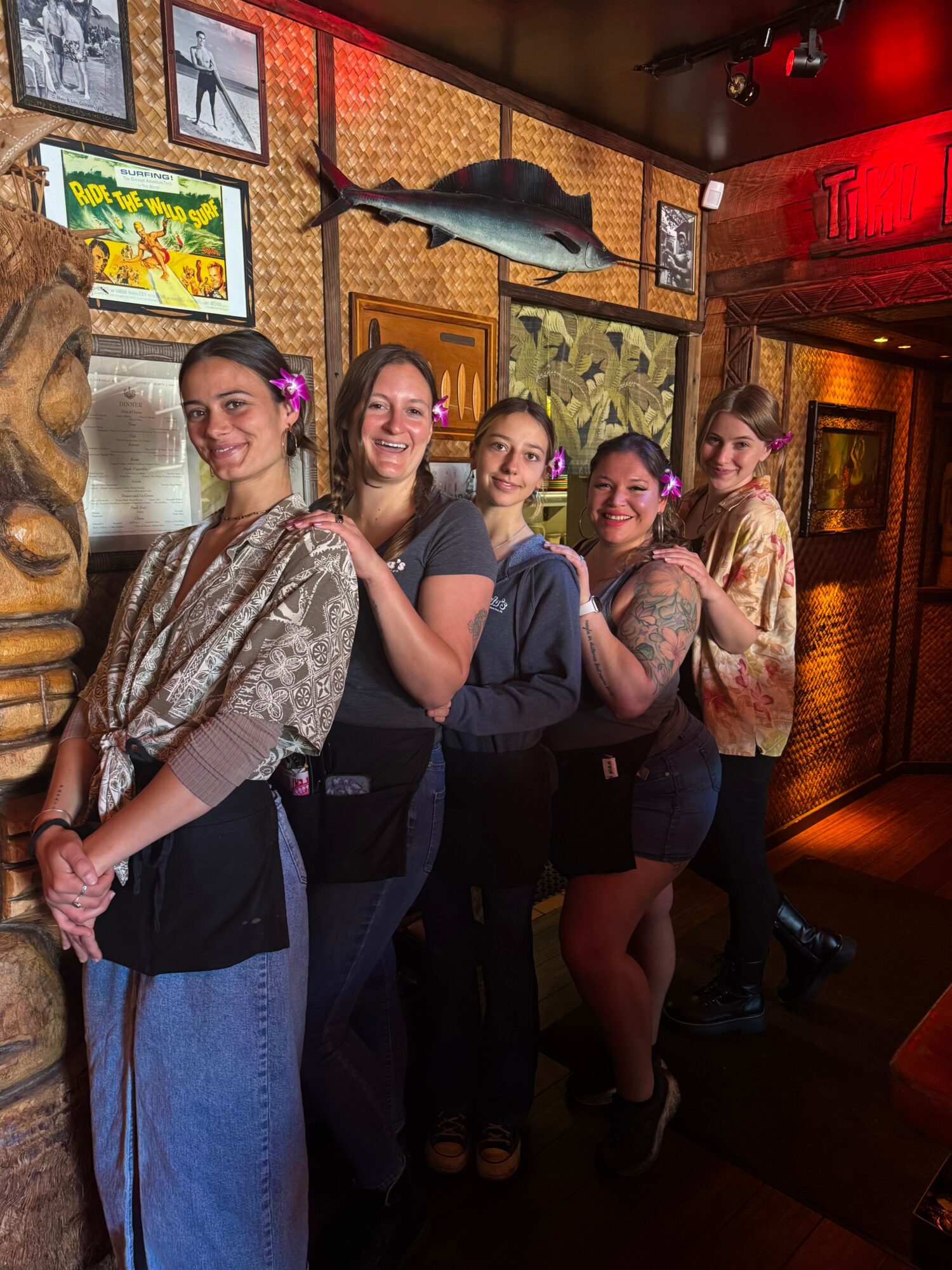 Five women stand in a line inside a restaurant, smiling, with colorful artwork and a fish decoration on the wall behind them.