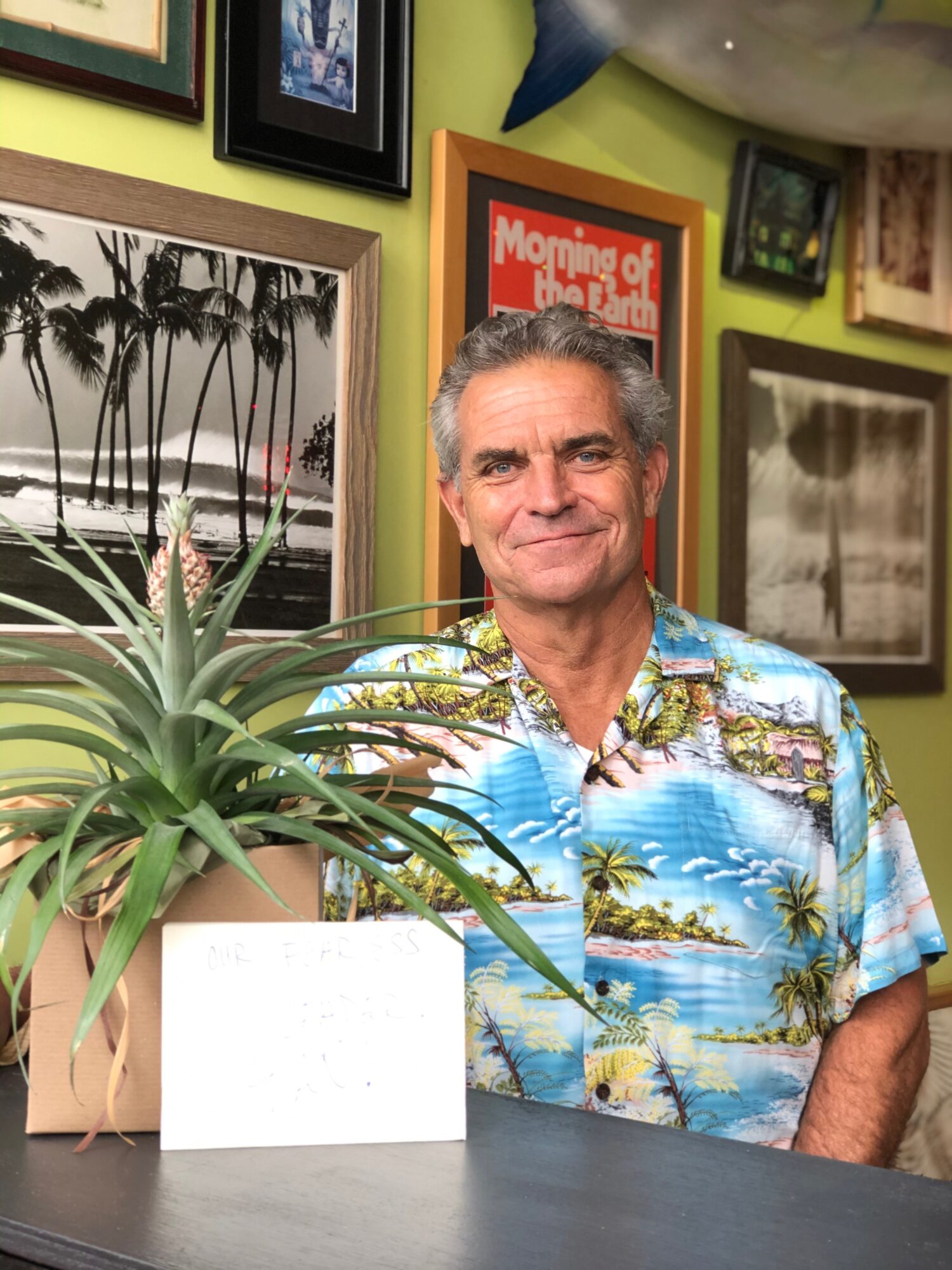 Man with gray hair wearing a colorful Hawaiian shirt, standing indoors near framed pictures and a potted plant.