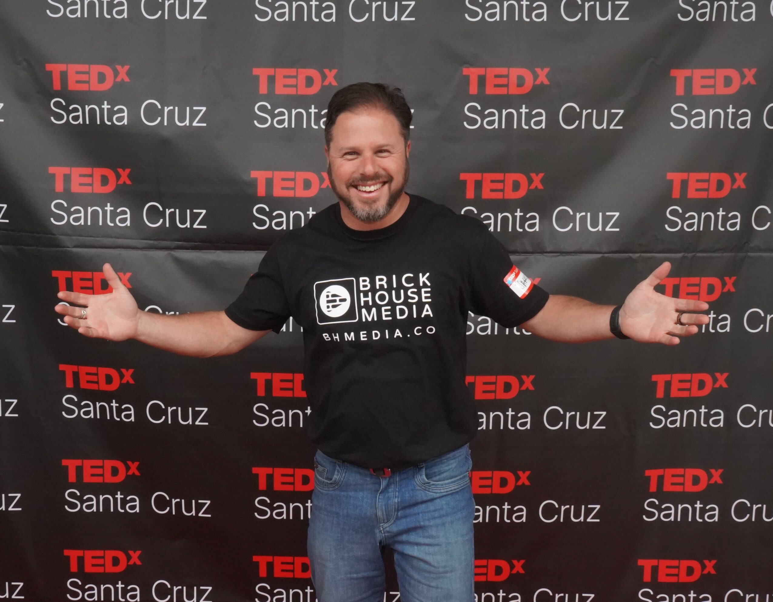 Man smiling with arms outstretched in front of a TEDx Santa Cruz backdrop, wearing a black T-shirt and jeans.