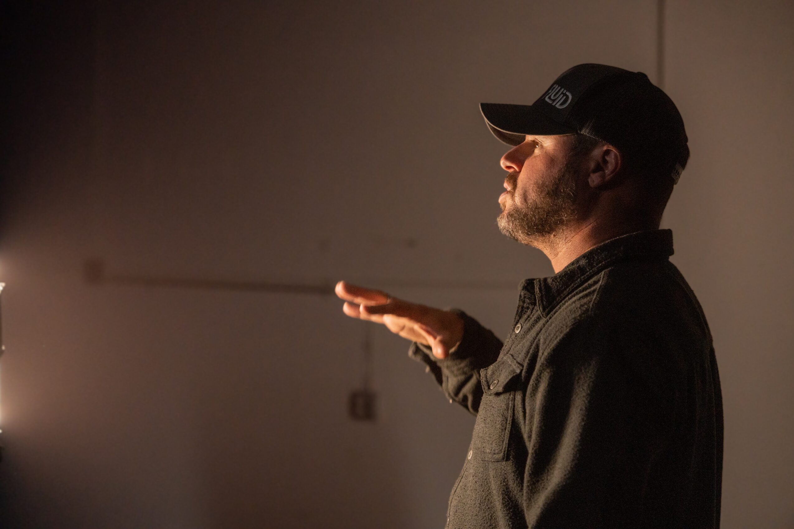 Man in a black cap and jacket faces right, with sunlight illuminating his face and hand, against a plain background.