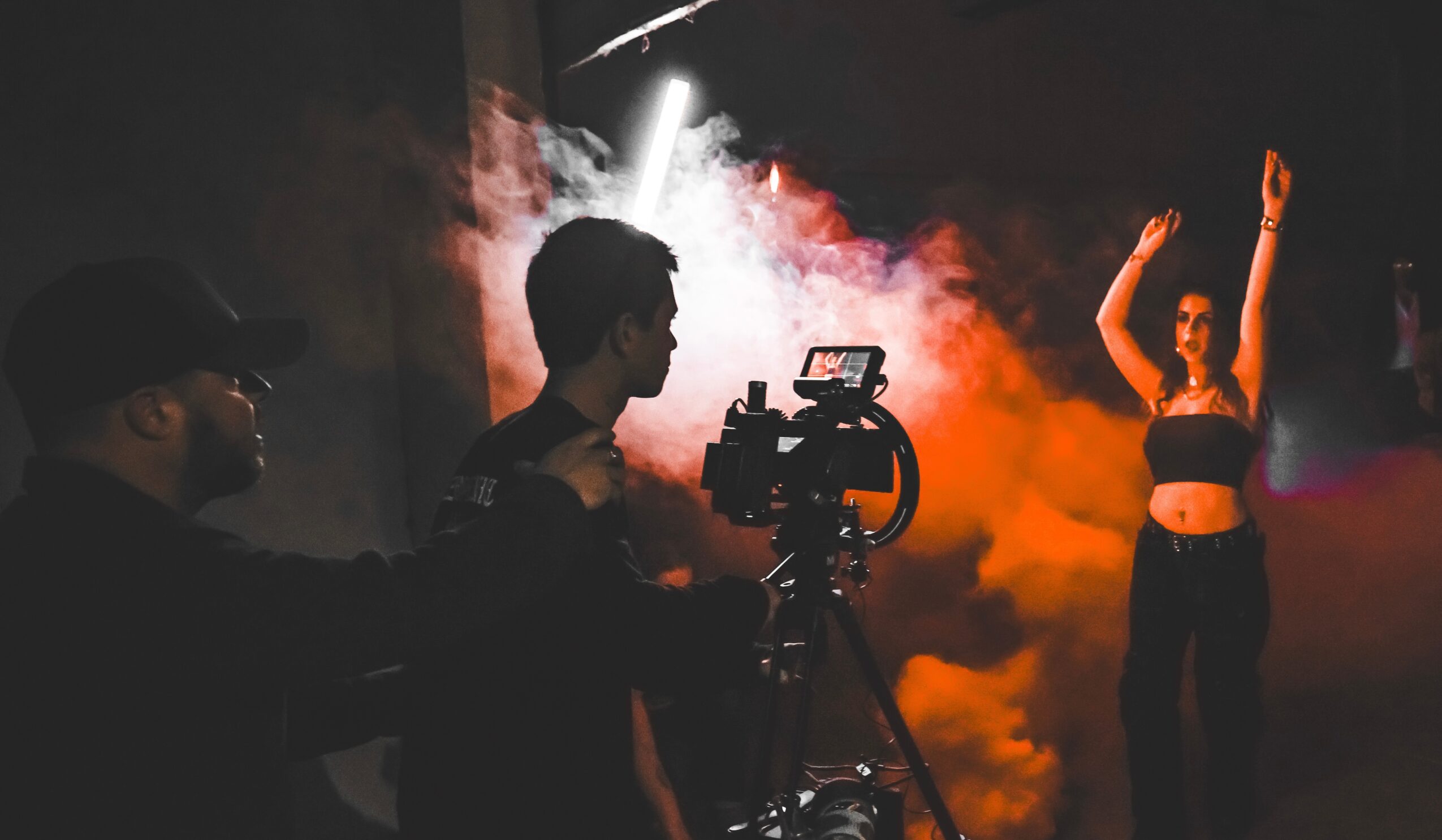 Person filming a dancer on stage with colorful smoke and lighting effects, dark background, two crew members visible.