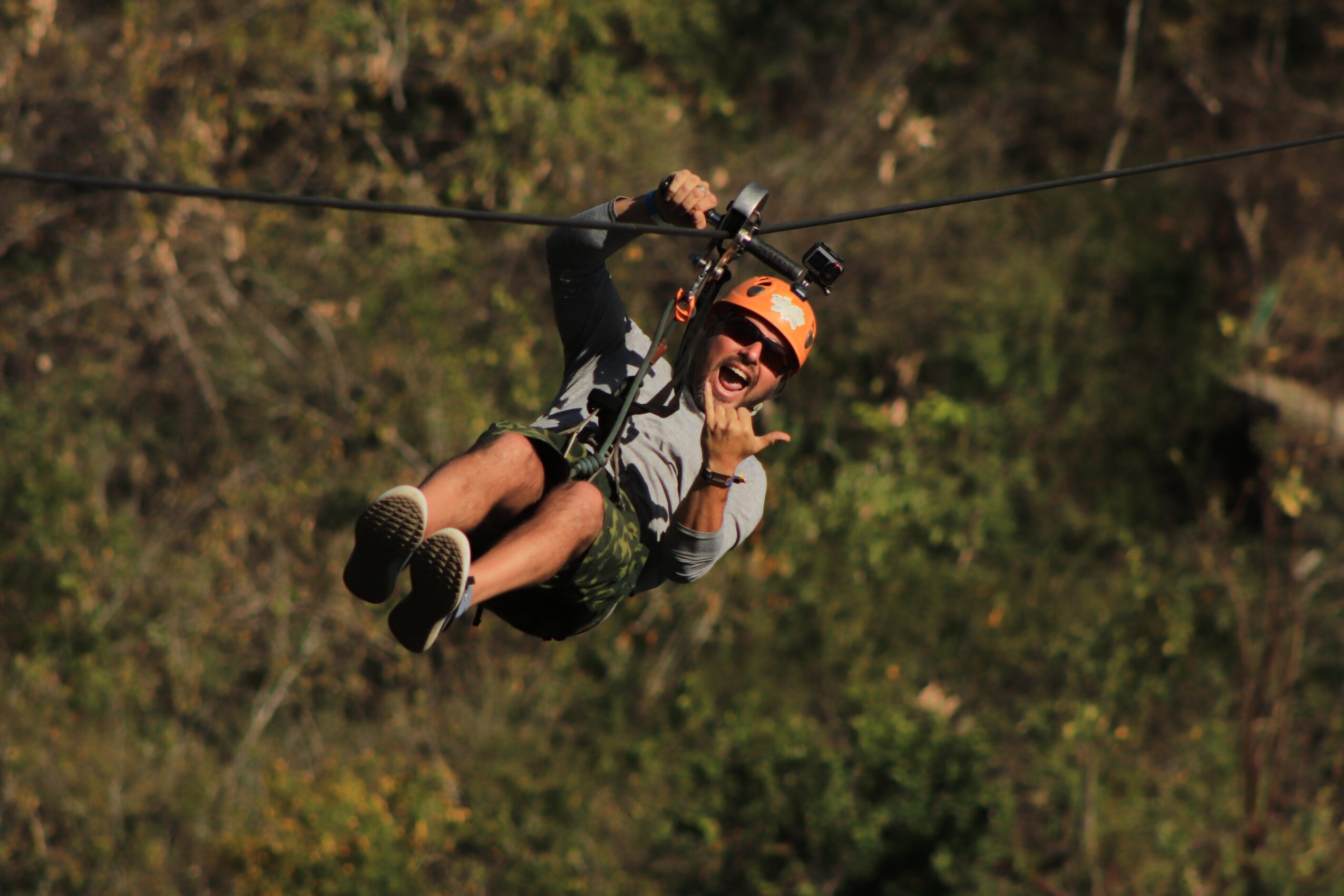 Person ziplining outdoors, wearing helmet and harness, smiling and talking on phone, trees in background.