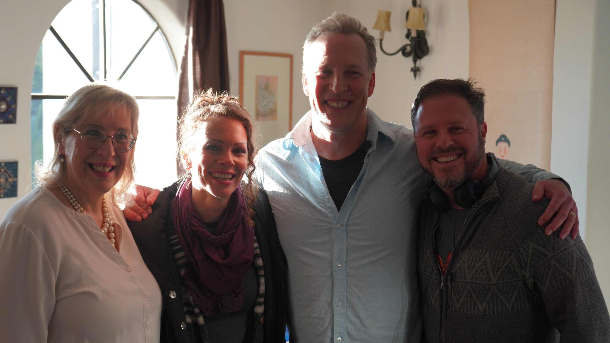 Four people smiling indoors, sunlight coming through a window, group close together, casual clothing, warm atmosphere.
