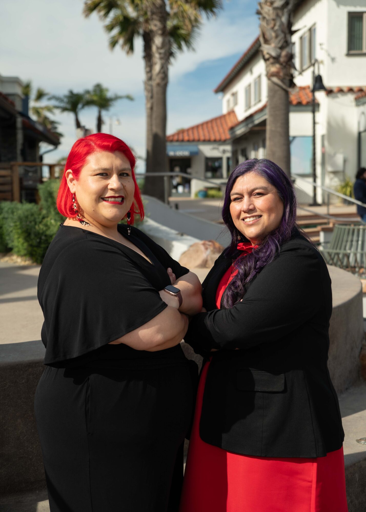 Two women with colorful hair smiling outdoors, palm trees and buildings in the background.
