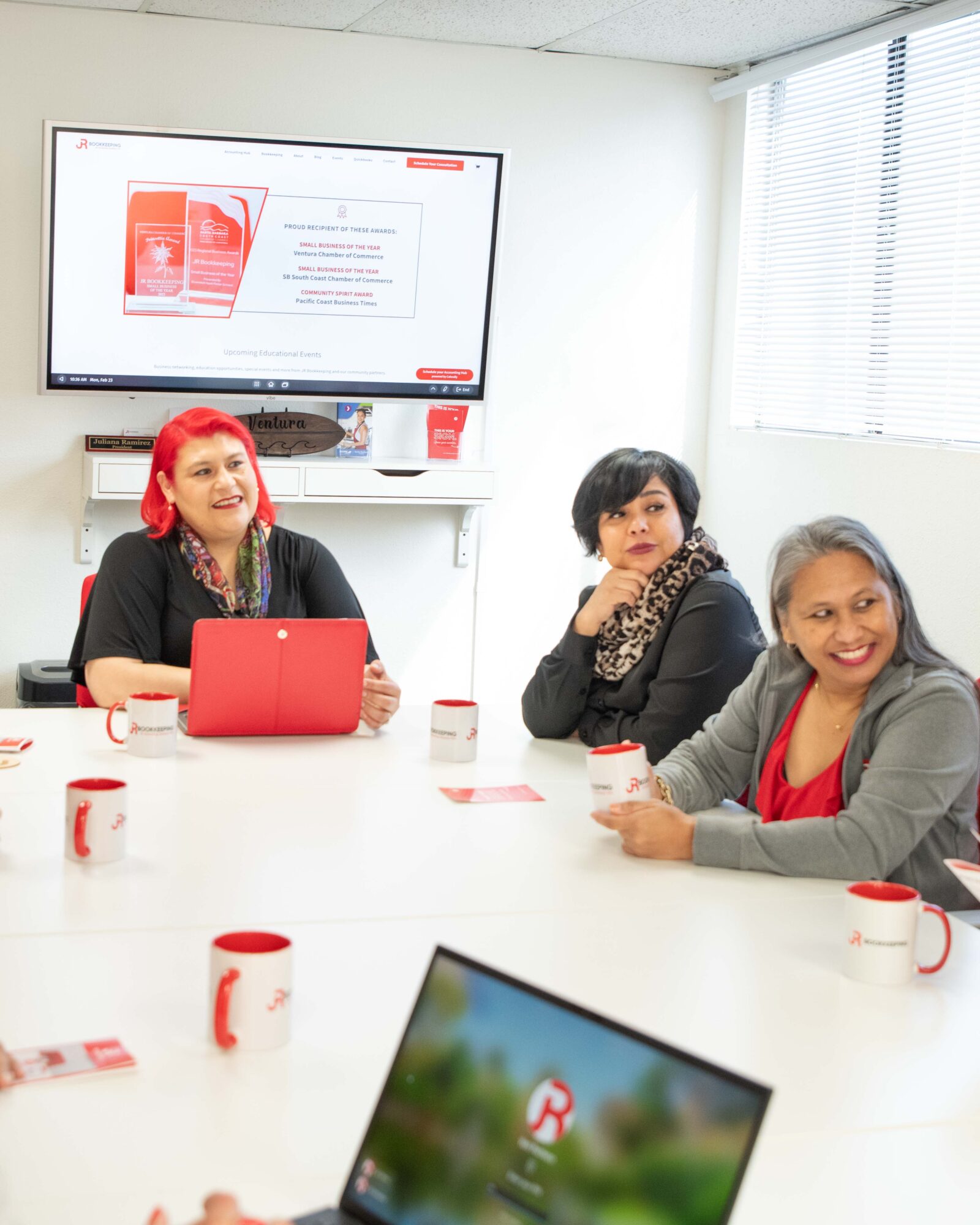 Three women sitting at a conference table with cups, a laptop, and a screen in the background, smiling and engaging.