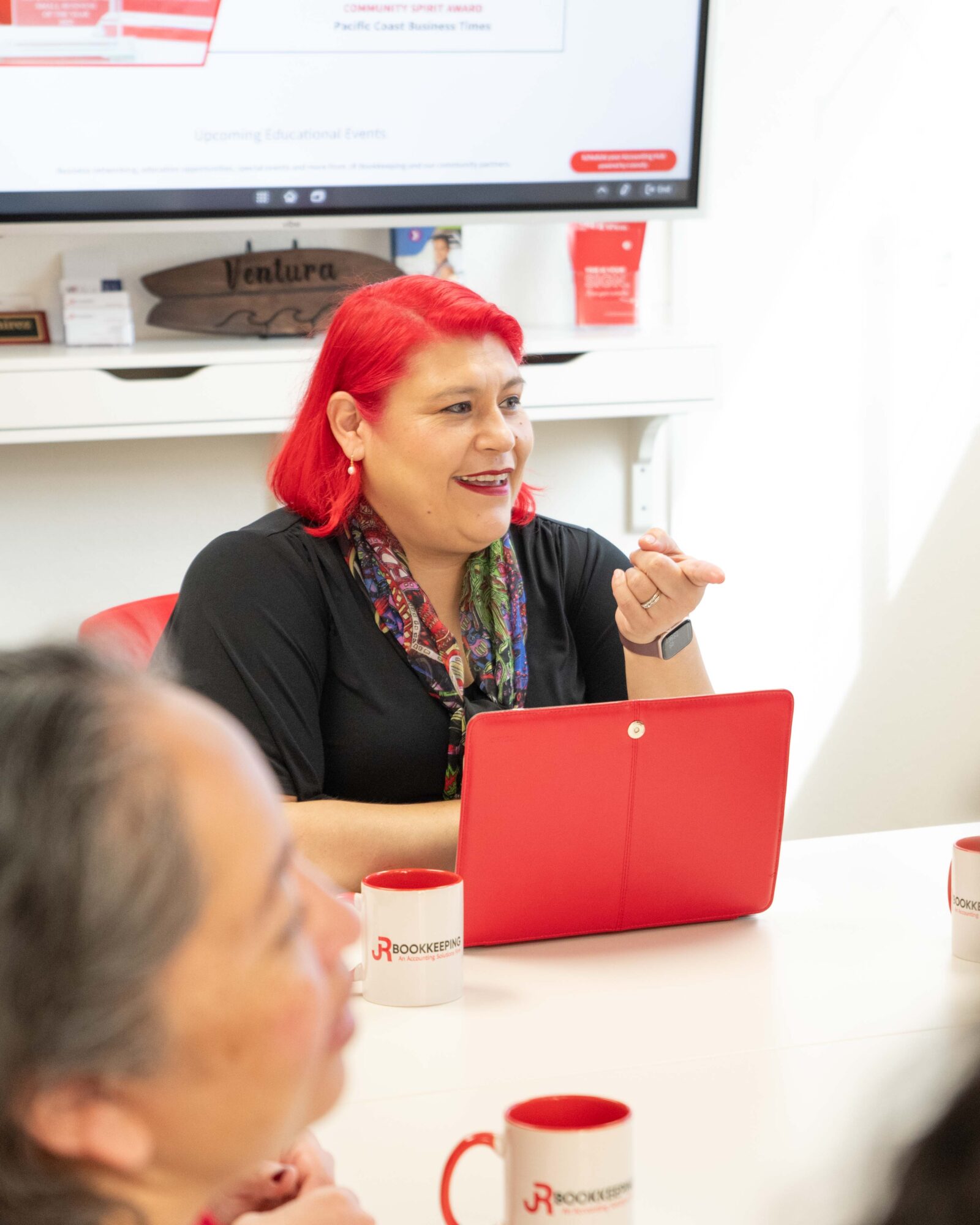 Woman with red hair speaking during a meeting, sitting at a table with a red laptop and mugs, in a bright room.