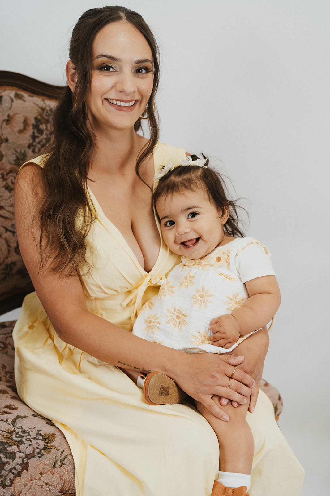 Woman and young girl sitting together, smiling, woman in yellow dress, girl in white dress with yellow pattern, holding hands.