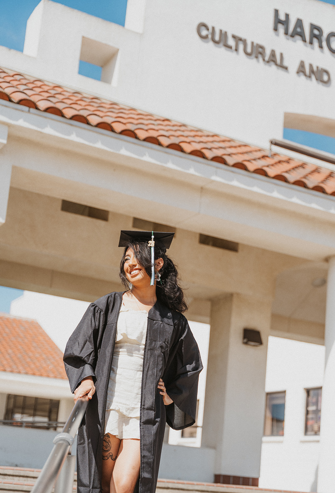 Person in graduation gown smiling outside a building with a sign reading 'Cultural and...'.