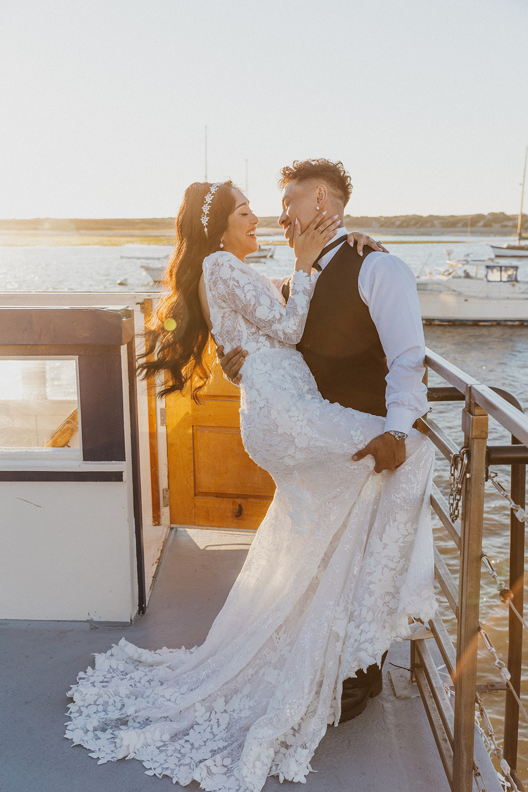A couple in wedding attire stands on a boat deck near water, embracing and looking at each other during sunset.