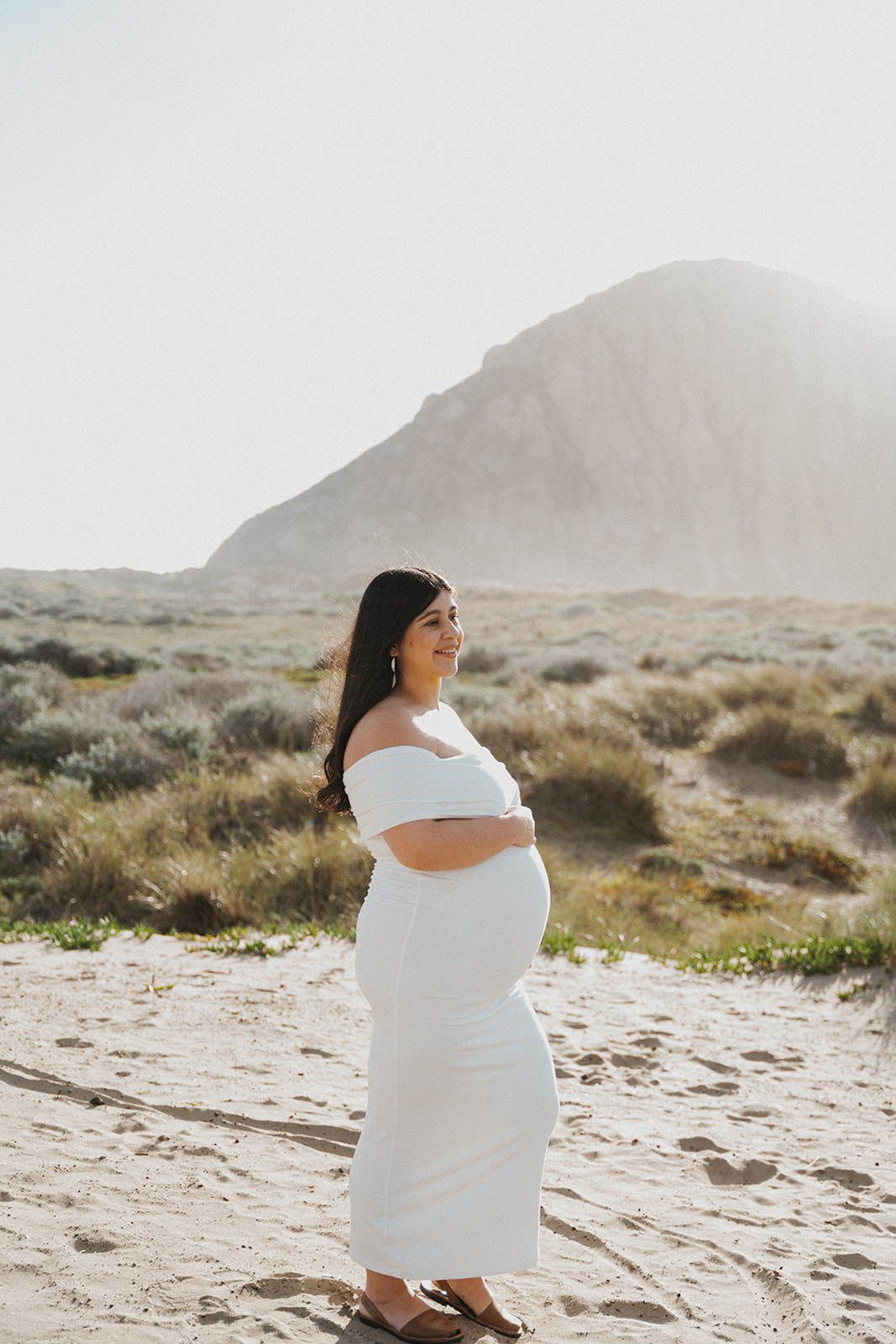 Pregnant woman in white dress standing outdoors with mountain and desert landscape in background.
