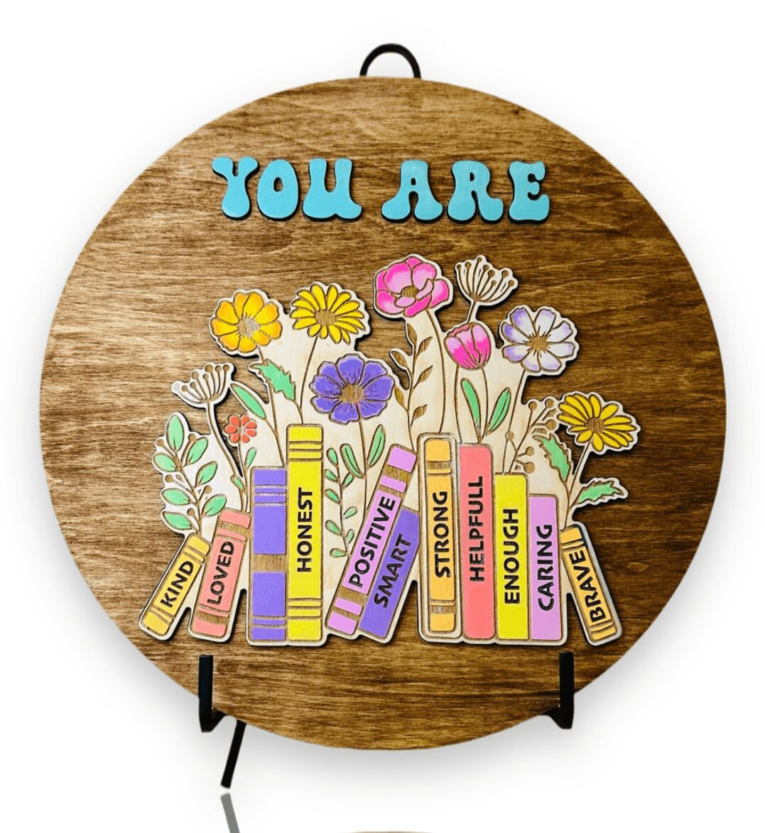Wooden circular plaque with colorful flowers and books, and the words 'You Are' at the top.