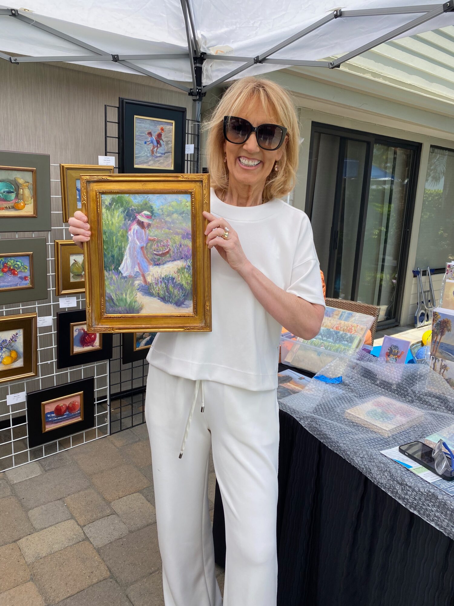 Woman with sunglasses holding a framed painting outdoors, surrounded by other artwork displayed on stands and tables.