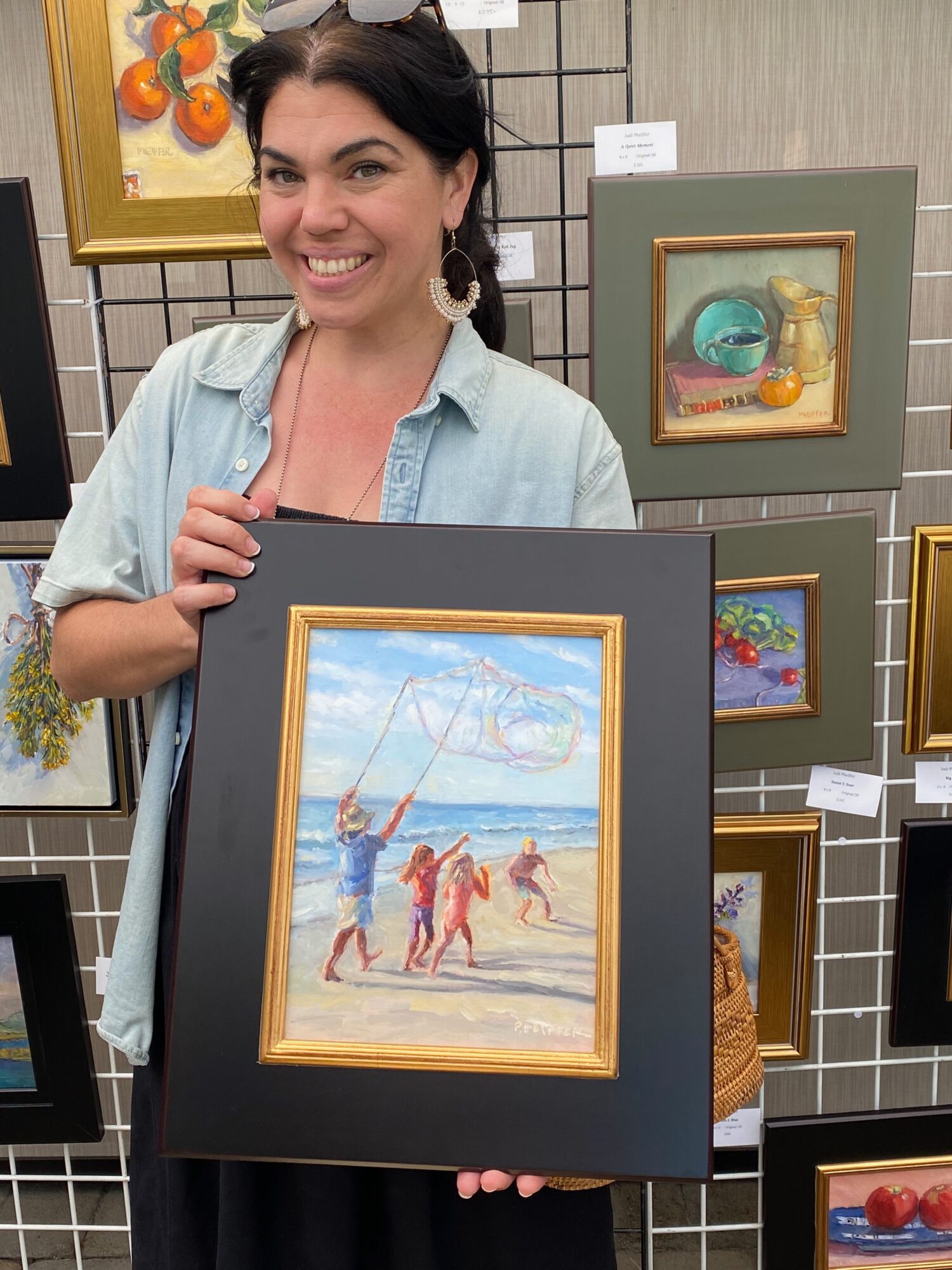 Woman holding framed beach scene painting, smiling, surrounded by other framed artworks on display.