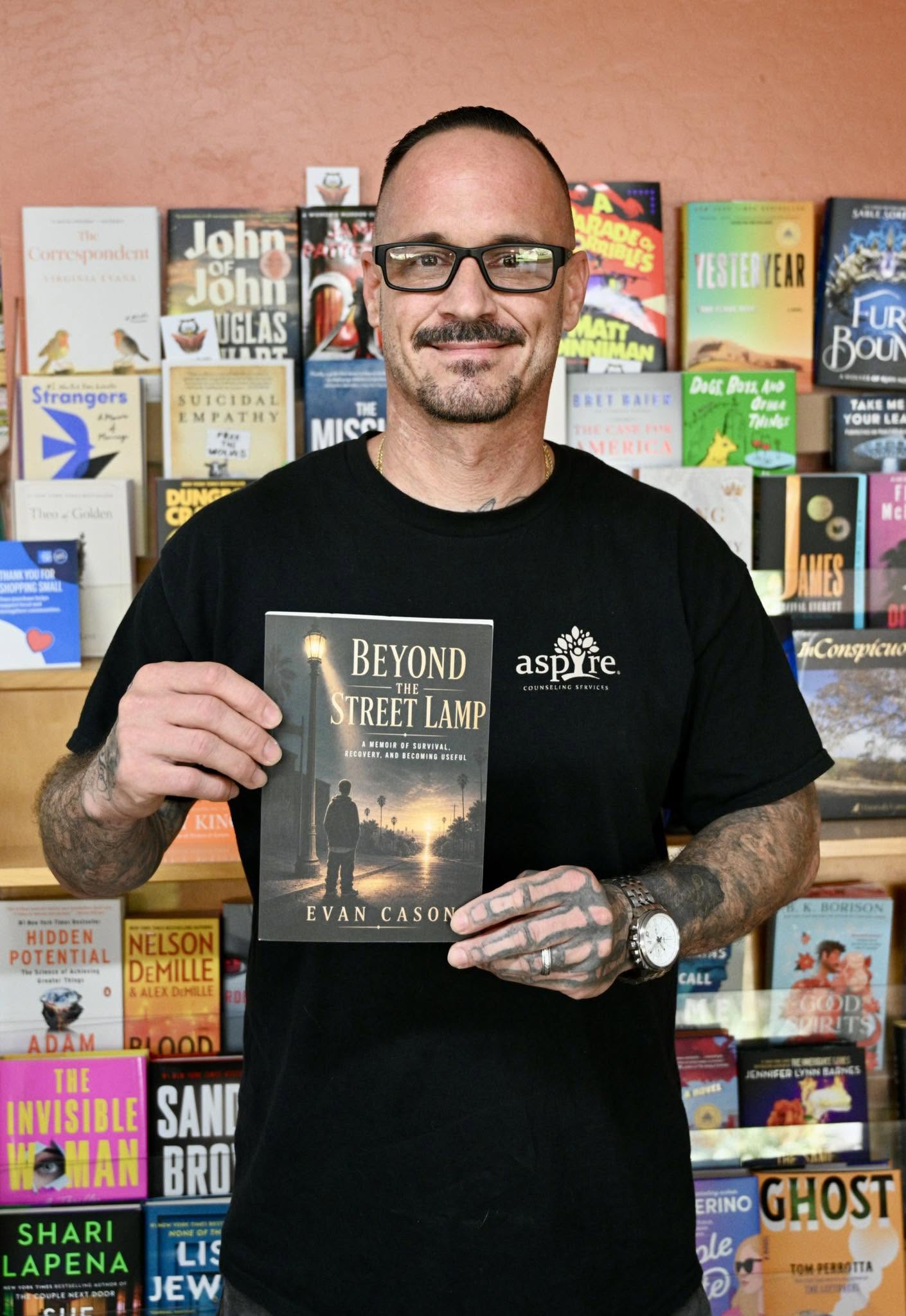 Man with glasses and tattoos holding a book in front of a colorful bookshelf, smiling, wearing a black shirt with a logo
