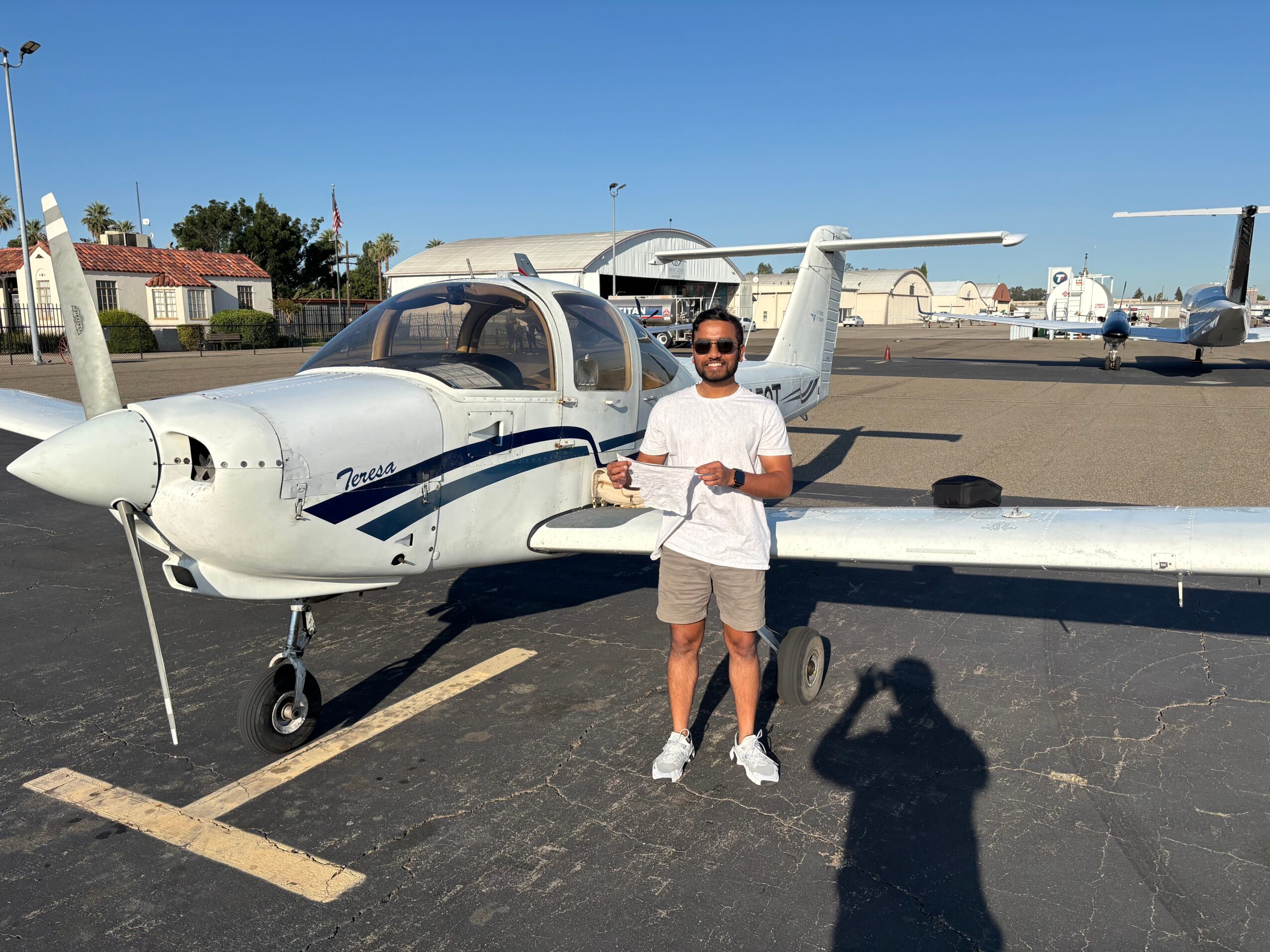 Person standing next to small private jet on tarmac, holding a drink, with clear blue sky in background.