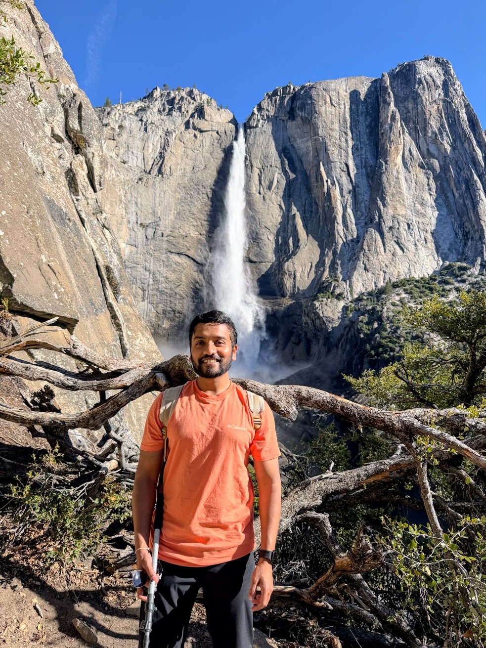 Man with beard and orange shirt standing outdoors in front of a waterfall and rocky cliffs under a blue sky.
