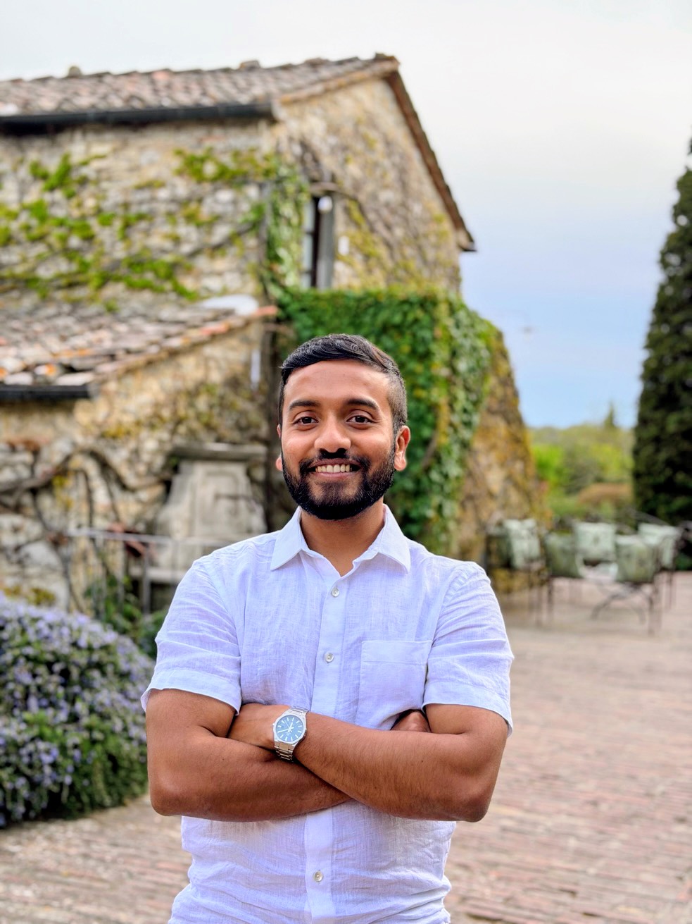 Man with crossed arms smiling outdoors in front of an old stone building and greenery