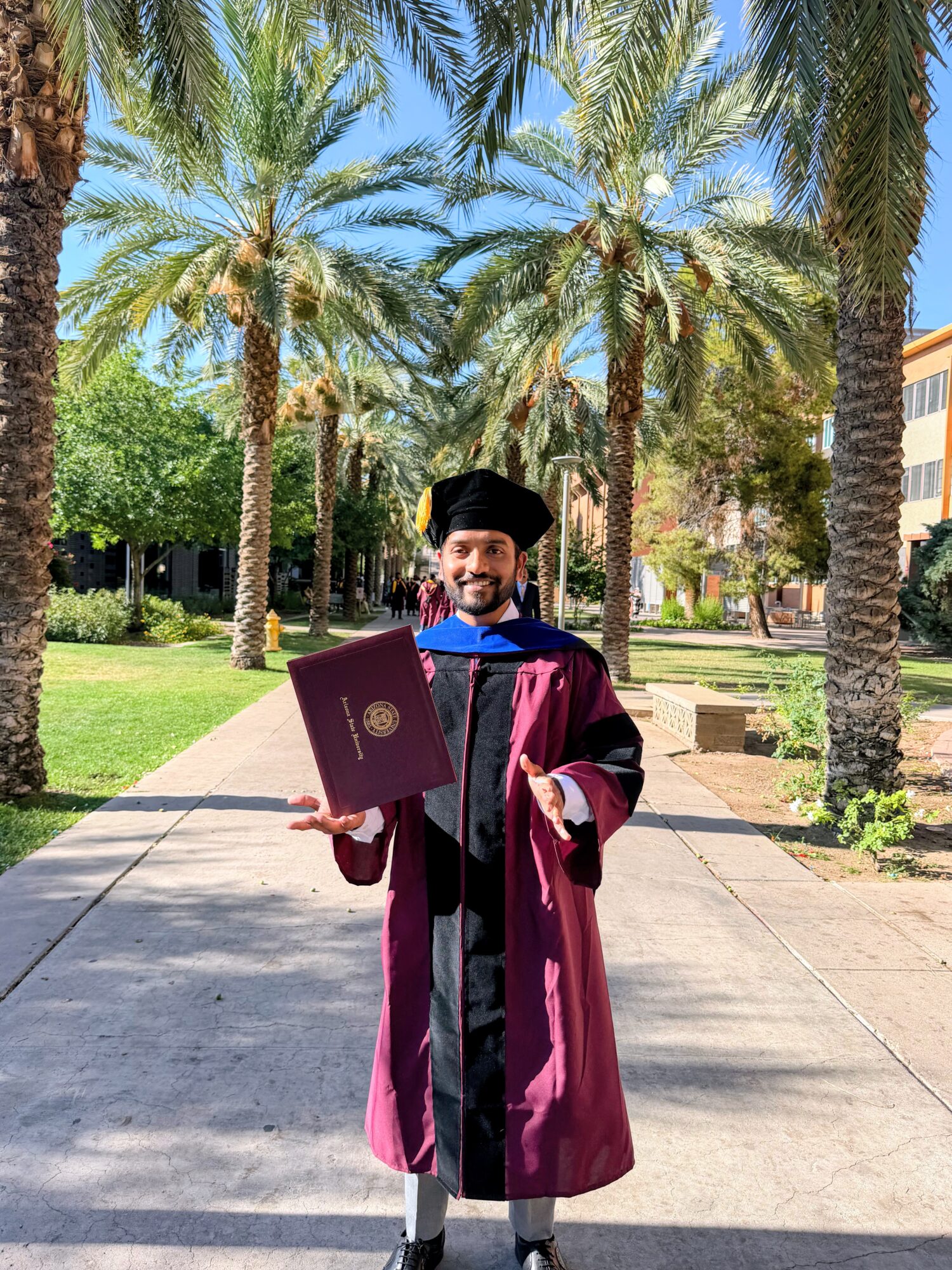 Graduate in cap and gown holding diploma, standing on pathway with palm trees and greenery in background.