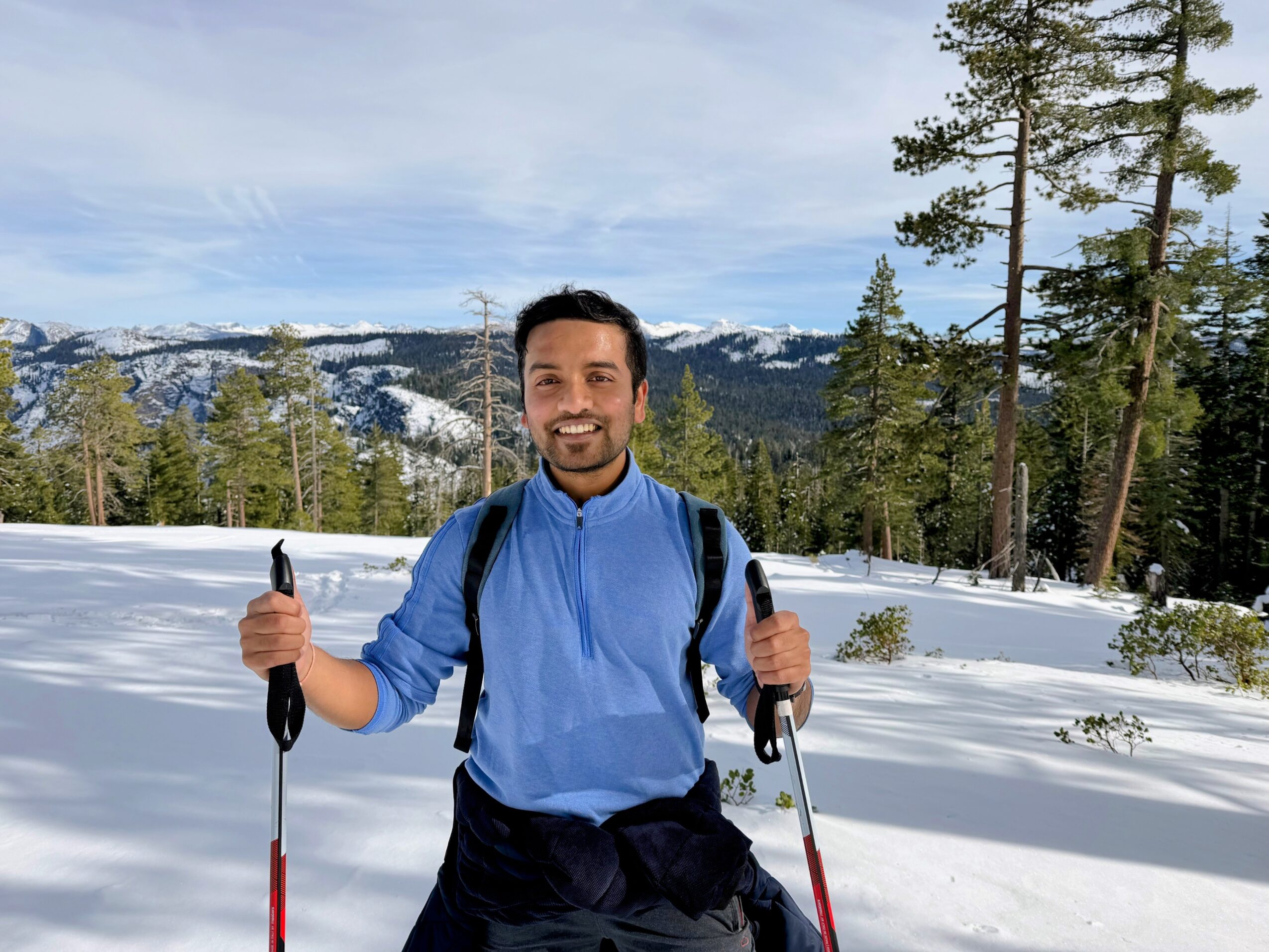 Man smiling outdoors in snowy landscape holding ski poles, wearing a blue jacket and backpack, with trees and mountains in background.