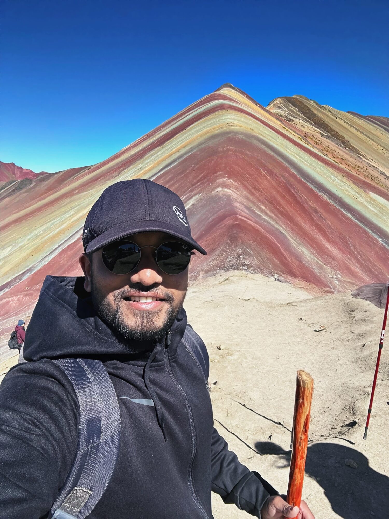 Man wearing sunglasses, cap, and backpack taking selfie in front of colorful layered mountain landscape.