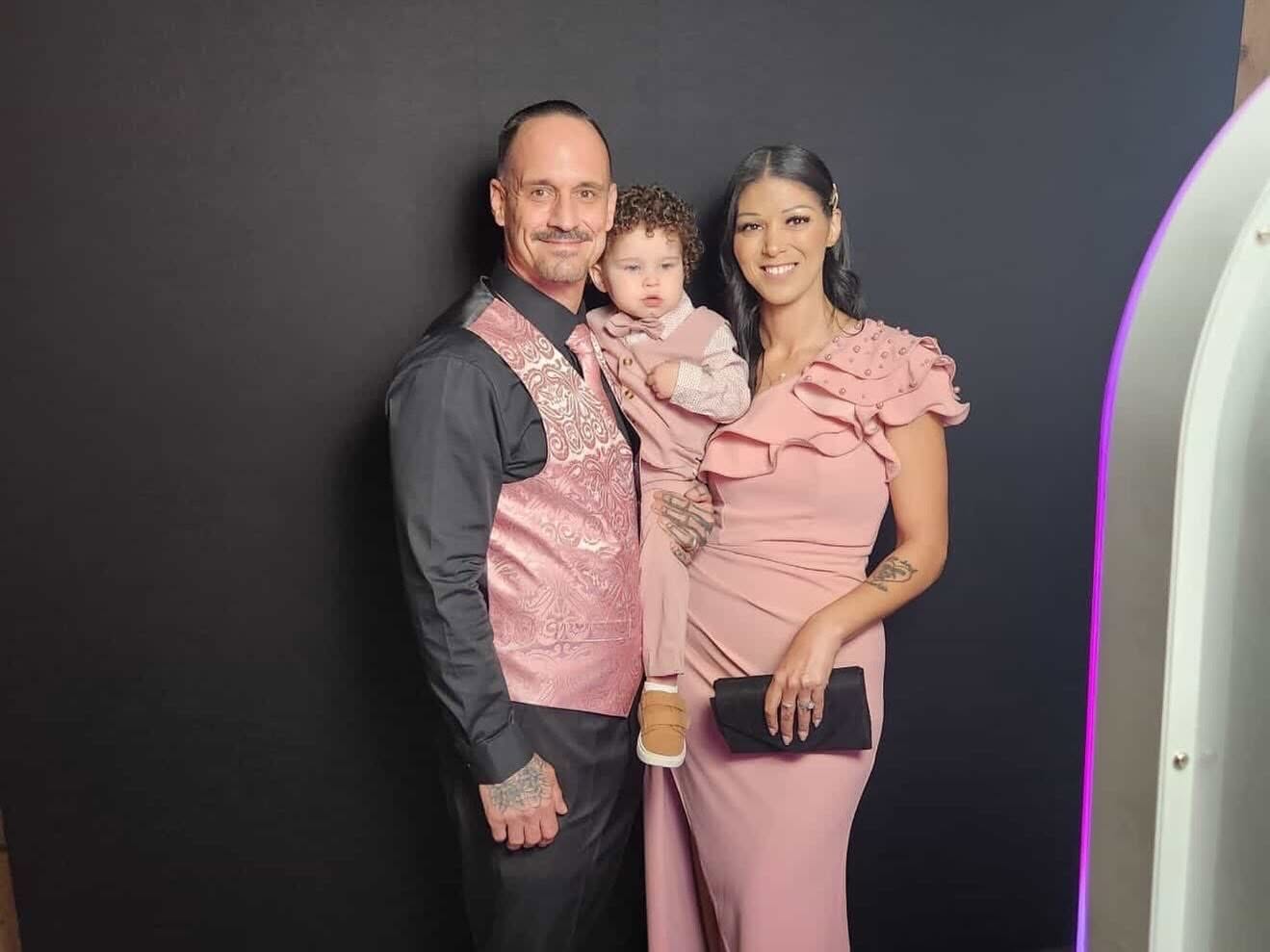 Family of three posing together, man and woman holding a child, all smiling, against a dark background.