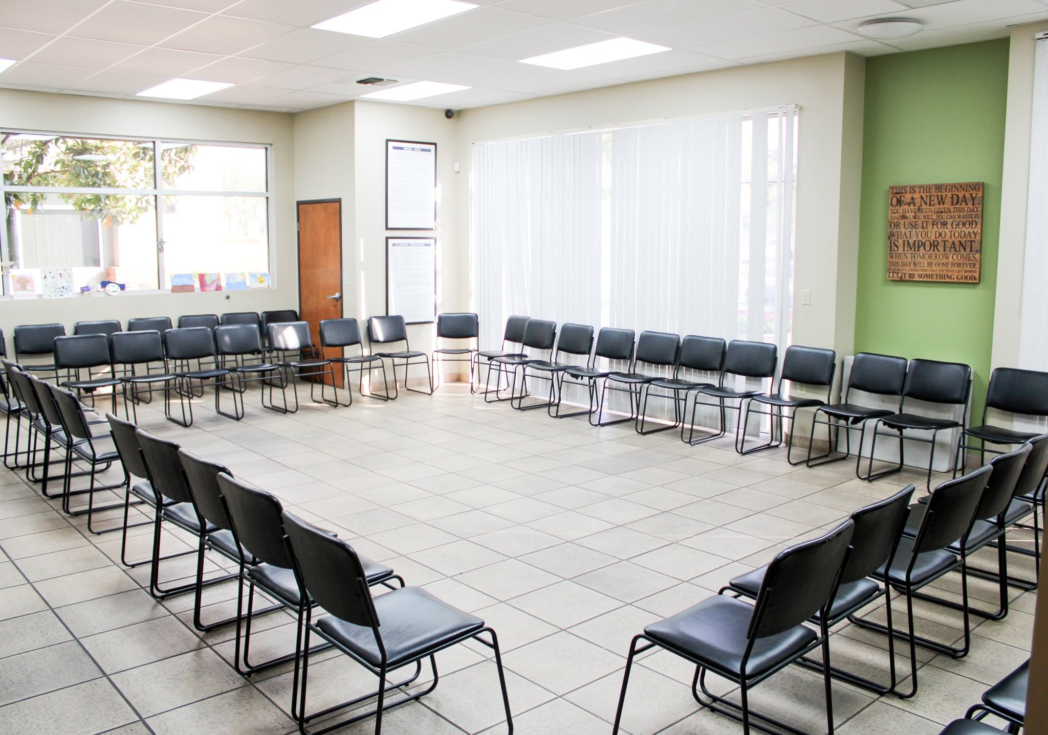 A large, empty room with chairs arranged in a square, white tiled floor, green and white walls, and large windows.
