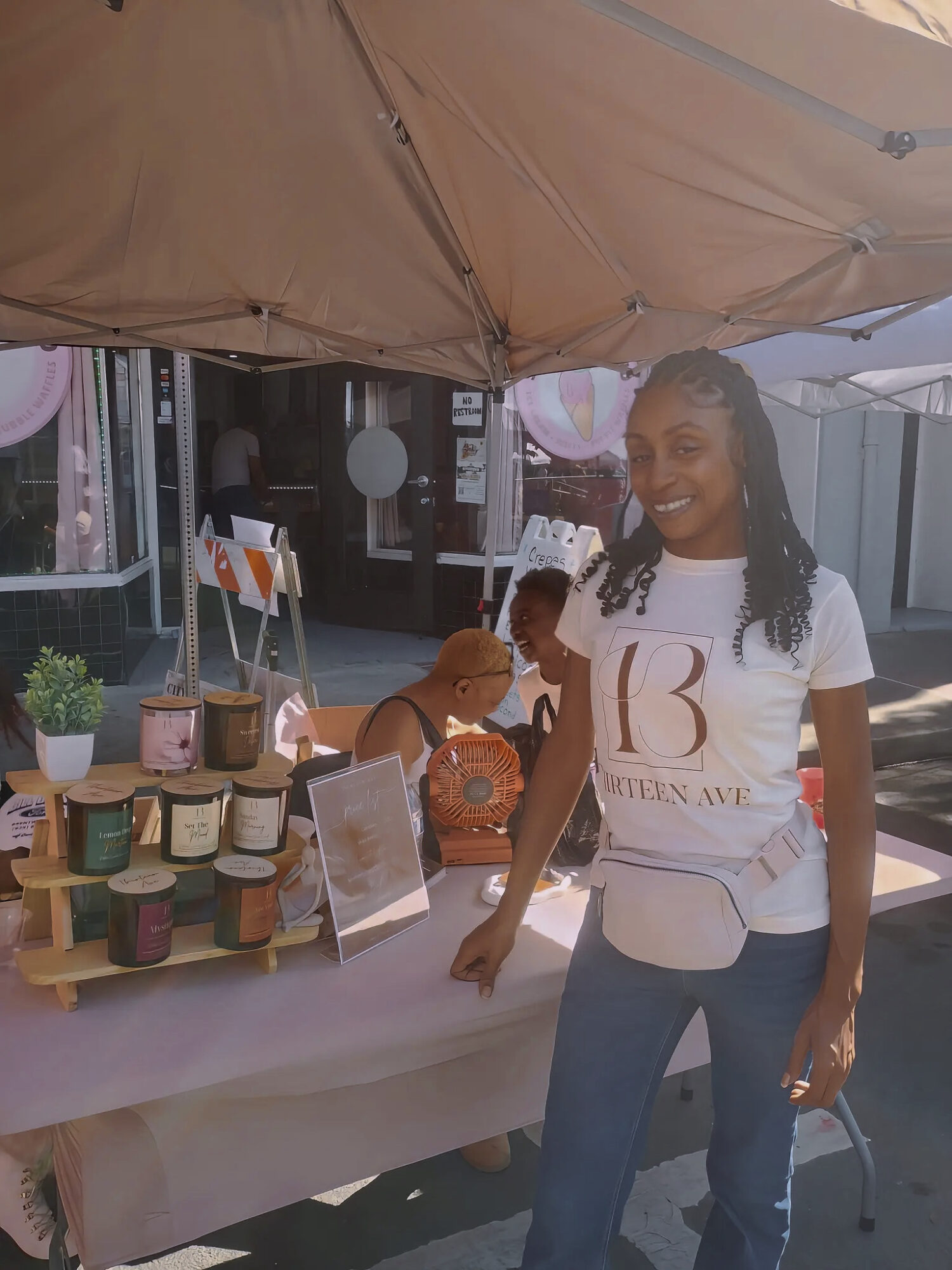 Woman smiling at an outdoor market stall with colorful jars and a table display, under a beige canopy.