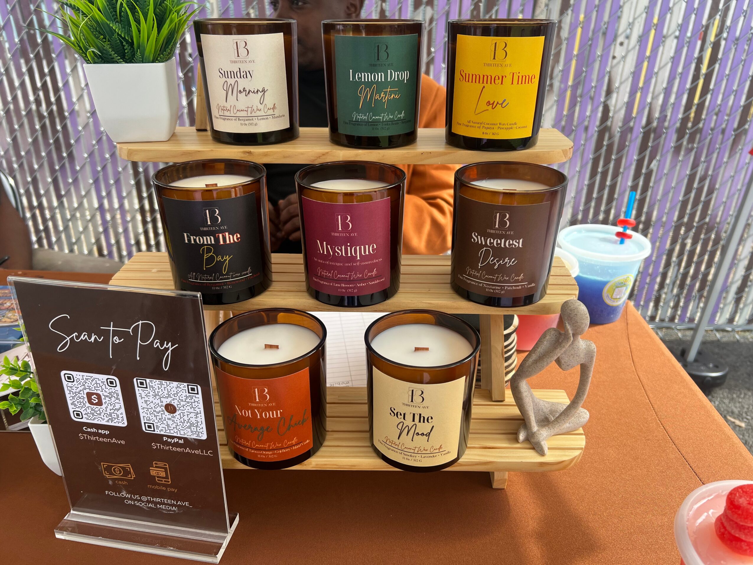 Display of nine candles on a wooden stand with a small gray sculpture, set on a table with a plant and a cup in the background.