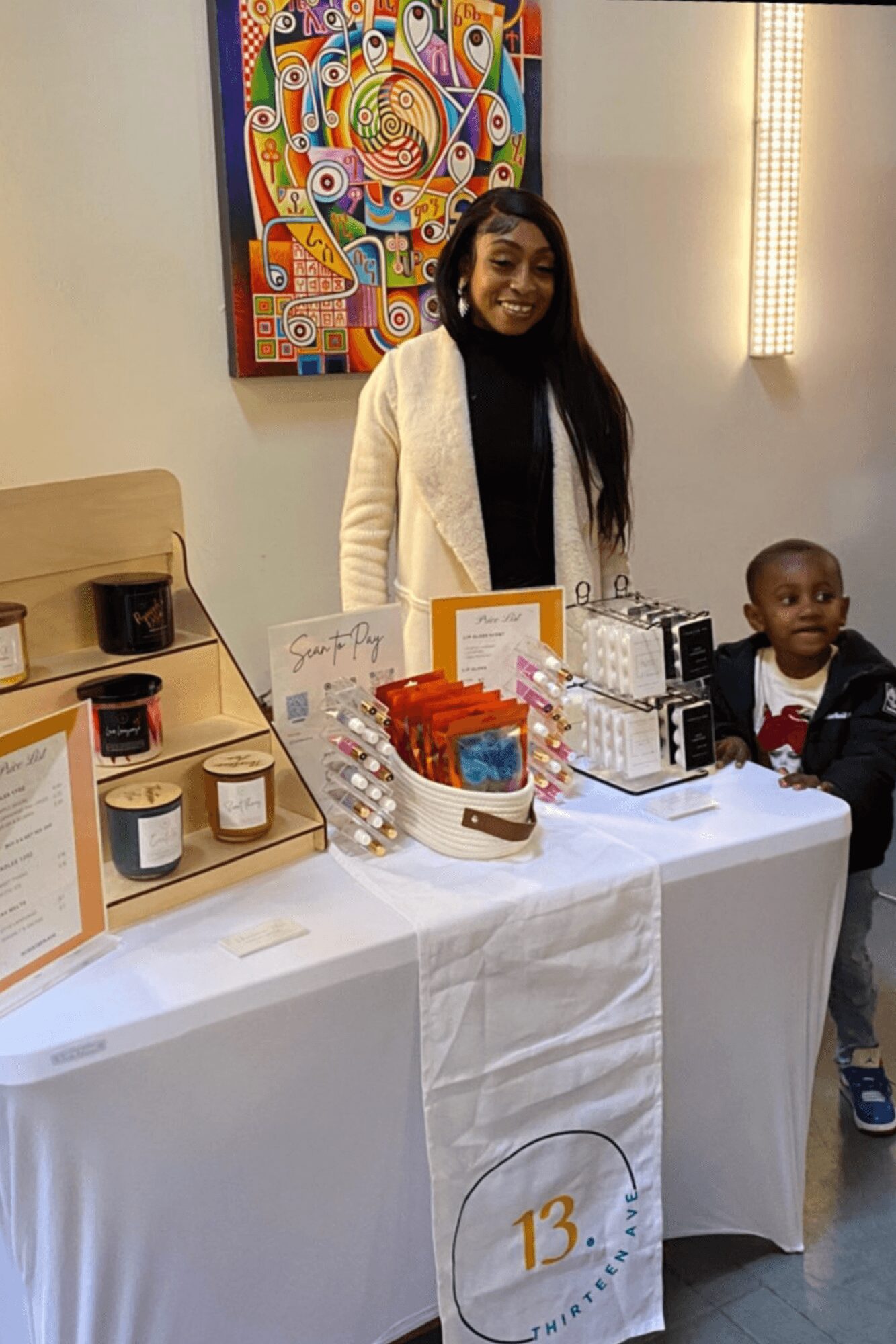 Woman standing behind a table with products, smiling, with a young boy nearby, in an indoor setting with colorful artwork on the wall.