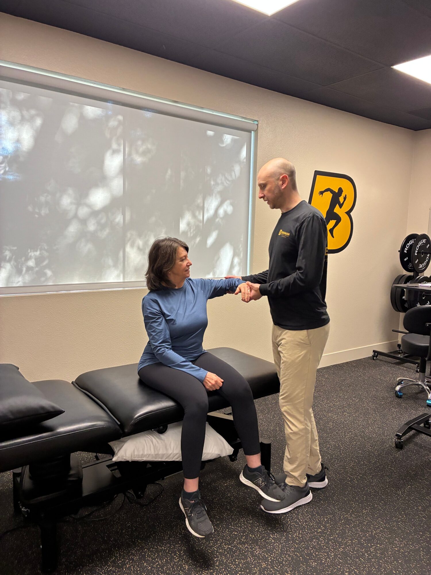 Two women shaking hands in a room with a large window, black chairs, and exercise equipment in the background.