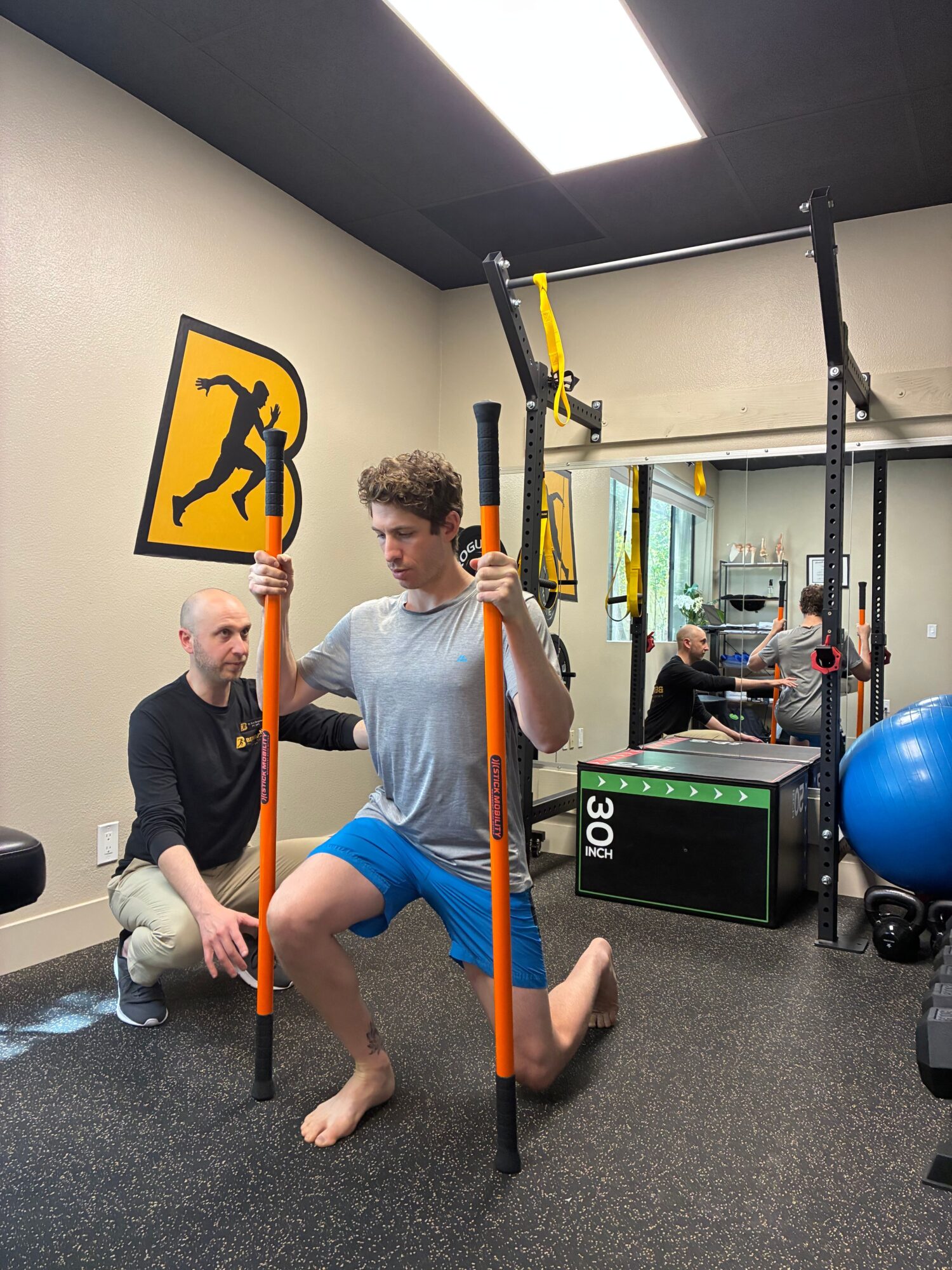 Two people in a gym with fitness equipment, one sitting on the floor and the other standing, holding poles, with a yellow sign on the wall.