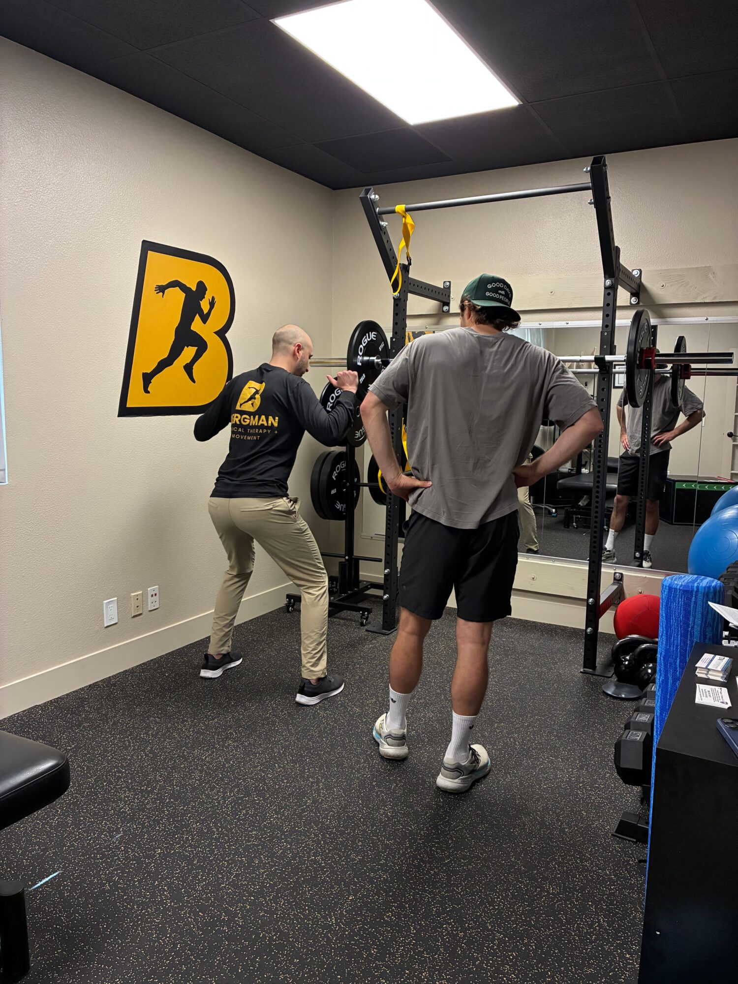 Two men in a gym, one with a backpack, standing near a yellow and black logo on the wall.
