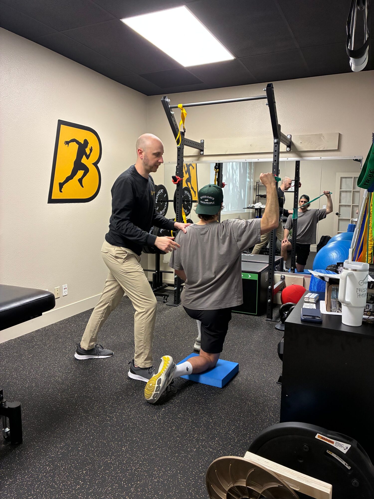 Two men in a gym, one standing and the other sitting on a blue exercise mat, with gym equipment around them.