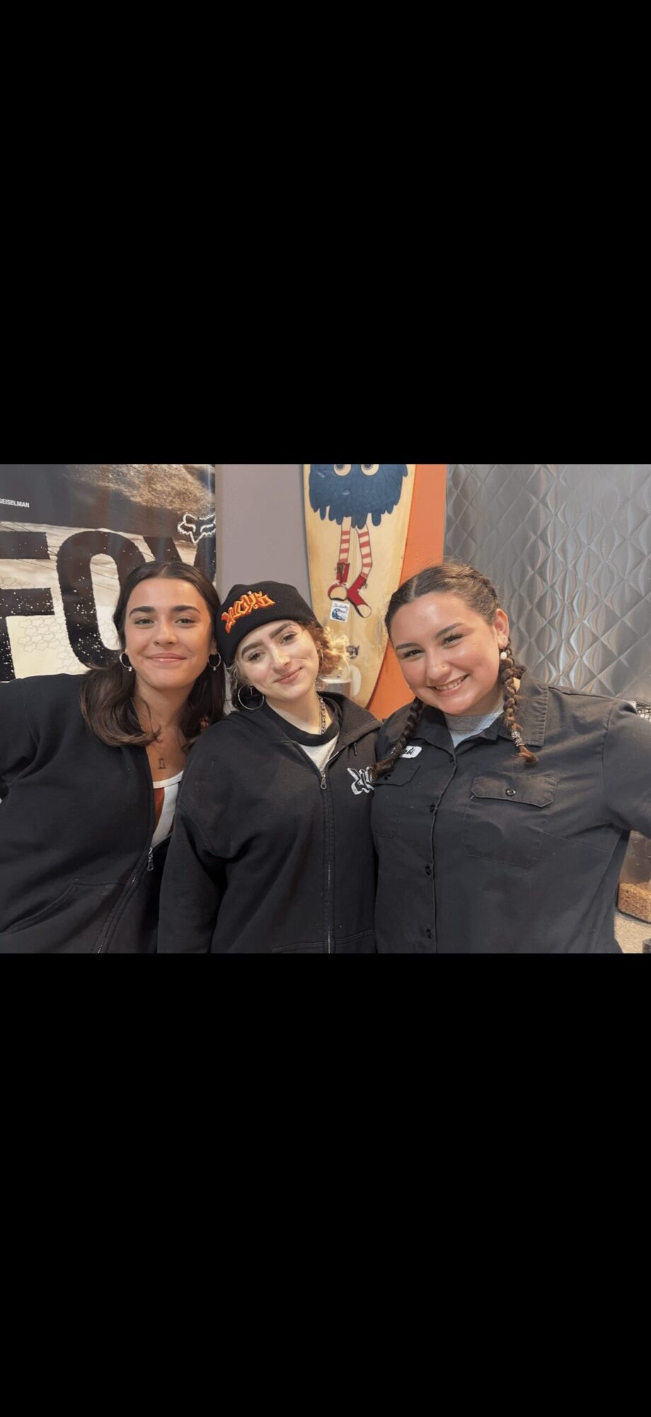 Three women standing together indoors, smiling, with colorful artwork in the background.