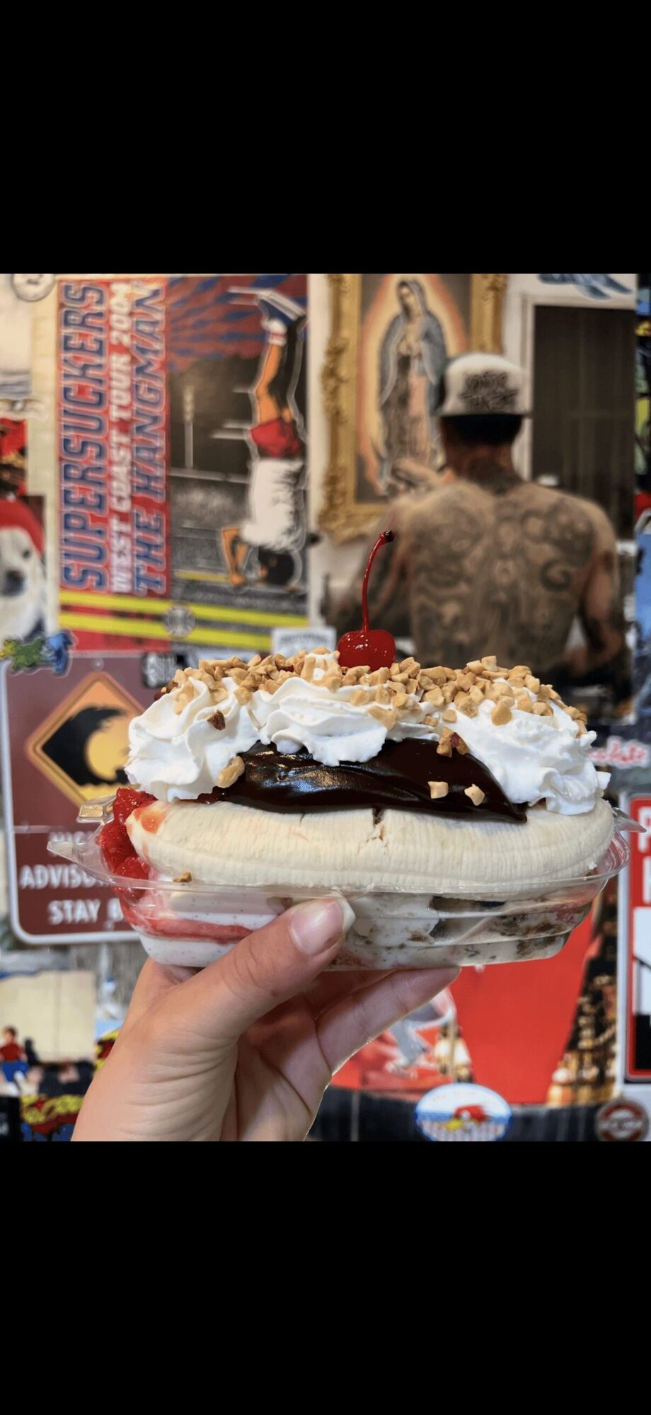Hand holding a dessert with whipped cream, chocolate, and banana slices in front of a colorful street scene with posters and a person in the background.