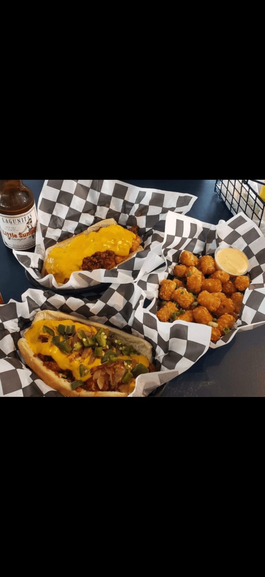 Two cheeseburgers with toppings and a side of fried chicken bites in checkered baskets, with a drink in the background.