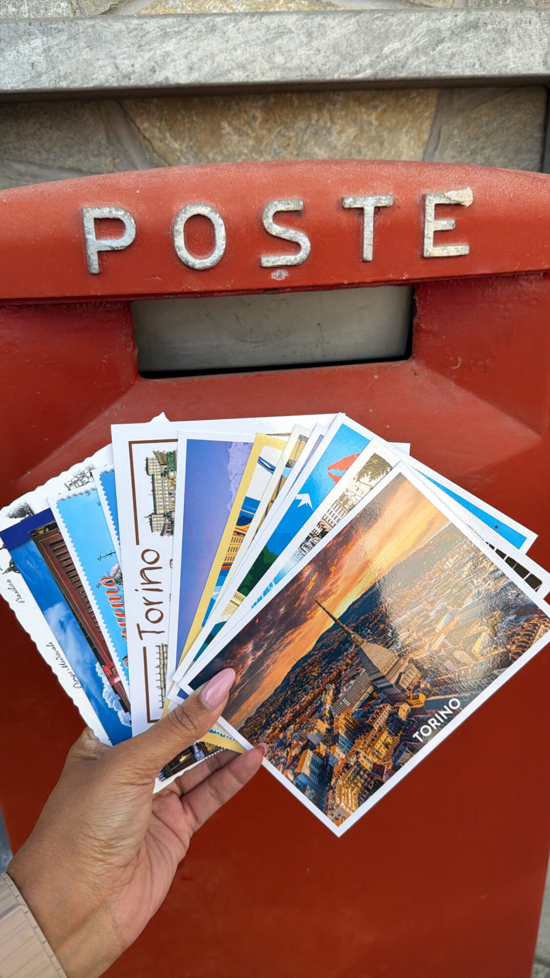 Hand holding postcards in front of a red mailbox with the word POSTE, various cityscapes and landscapes visible on postcards.