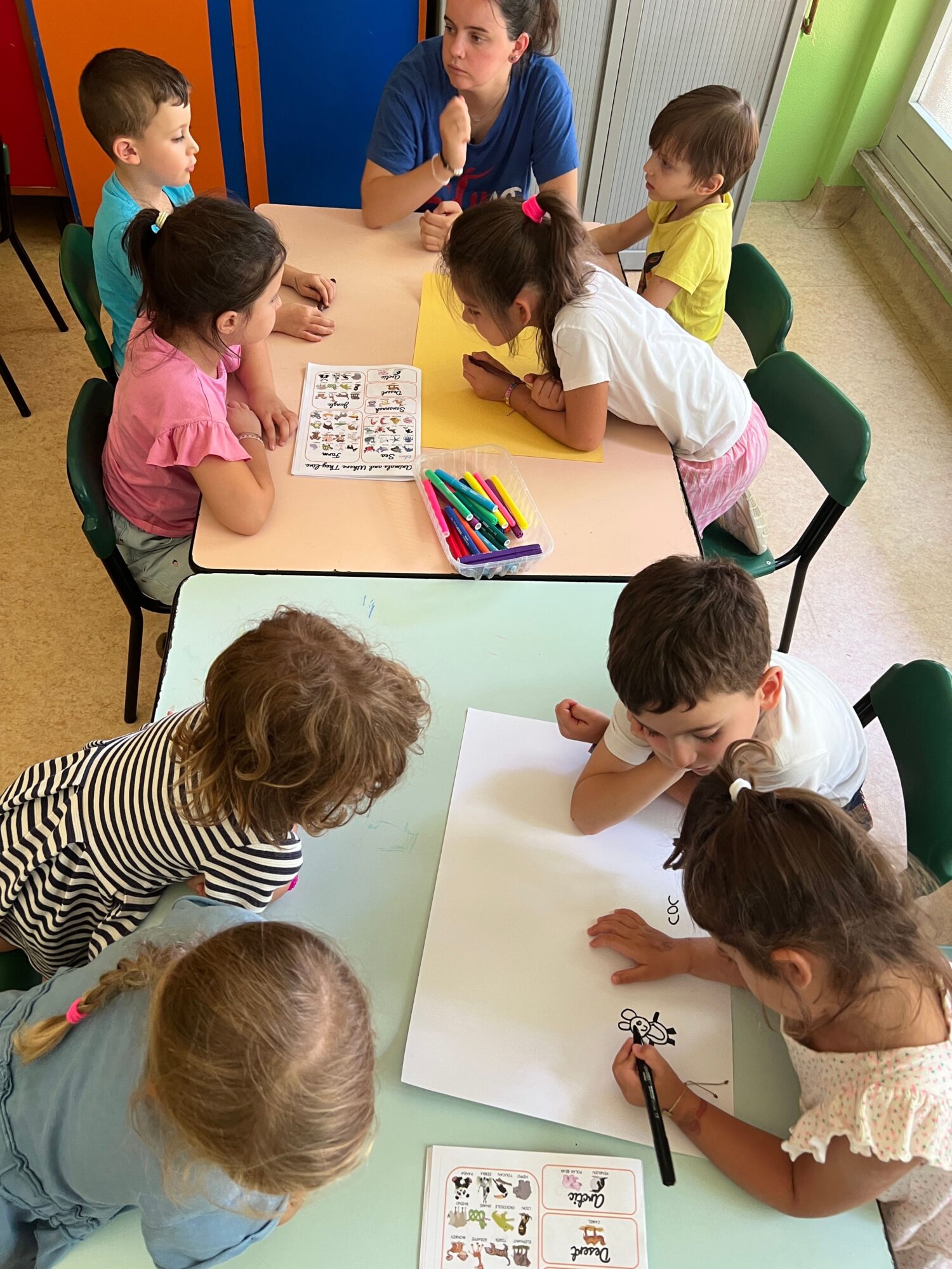 Children gather around a table drawing and coloring with markers, papers, and coloring books in a classroom.